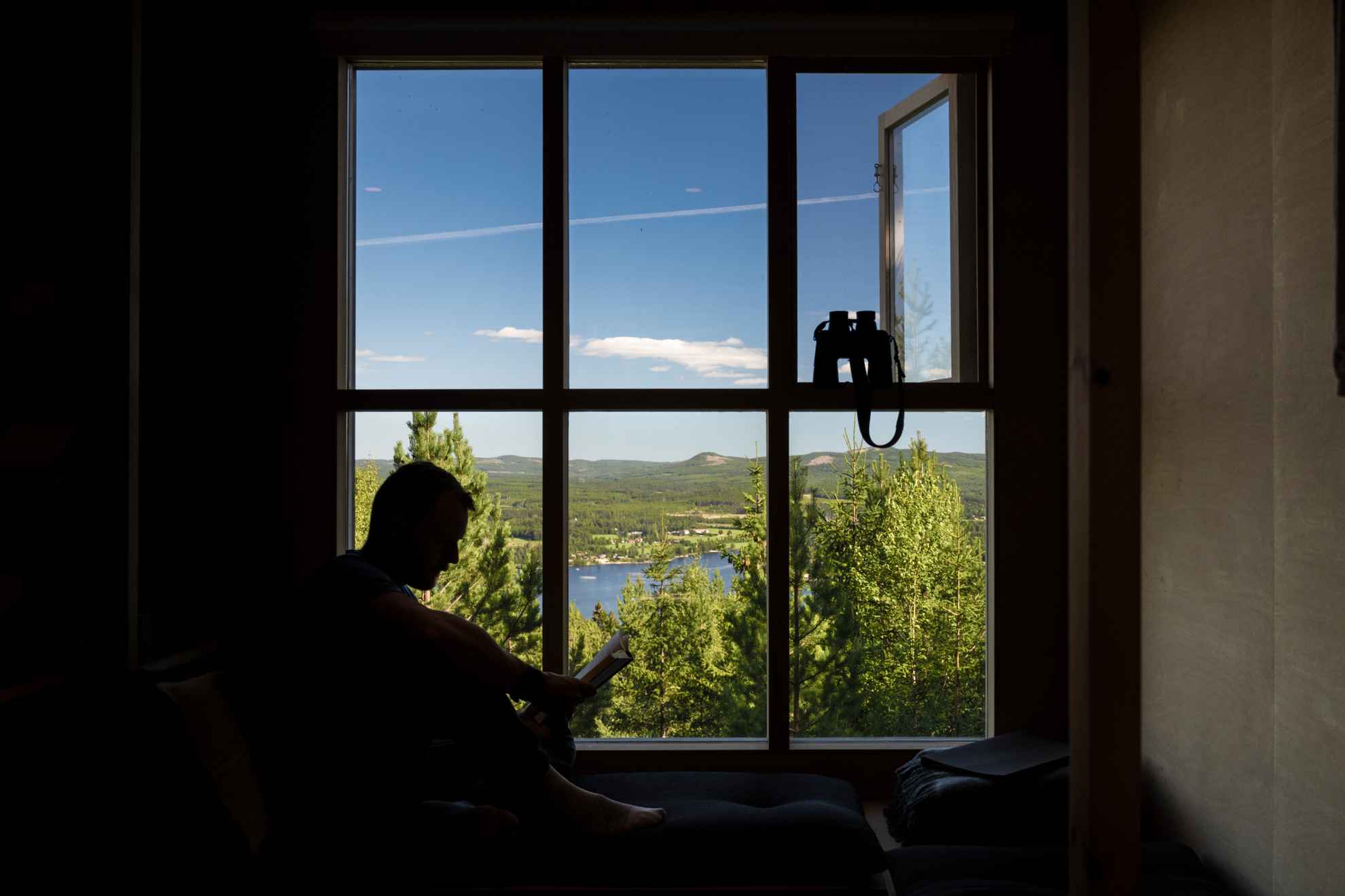 Silhouet van een man die een boek leest tegen een groot raam en uitkijkt over de blauwe lucht en de bergen erboven in Hälsingland, met een verrekijker in de aanslag.