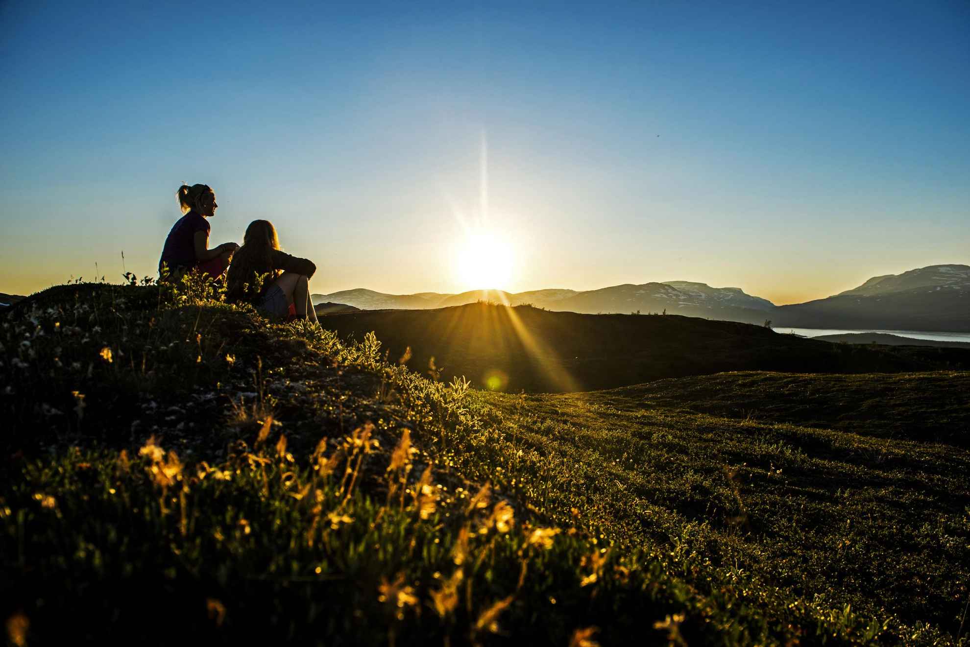Twee vrouwen zitten op een heuvel in de bergen en genieten van de middernachtzon.