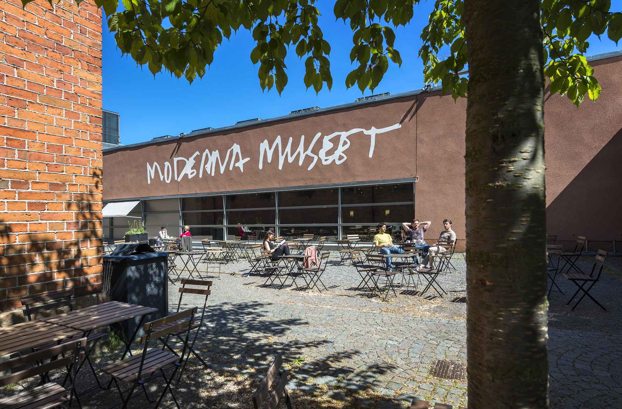Een binnenplaats met mensen die aan cafétafels zitten, buiten een gebouw met de tekst "Moderna museet".