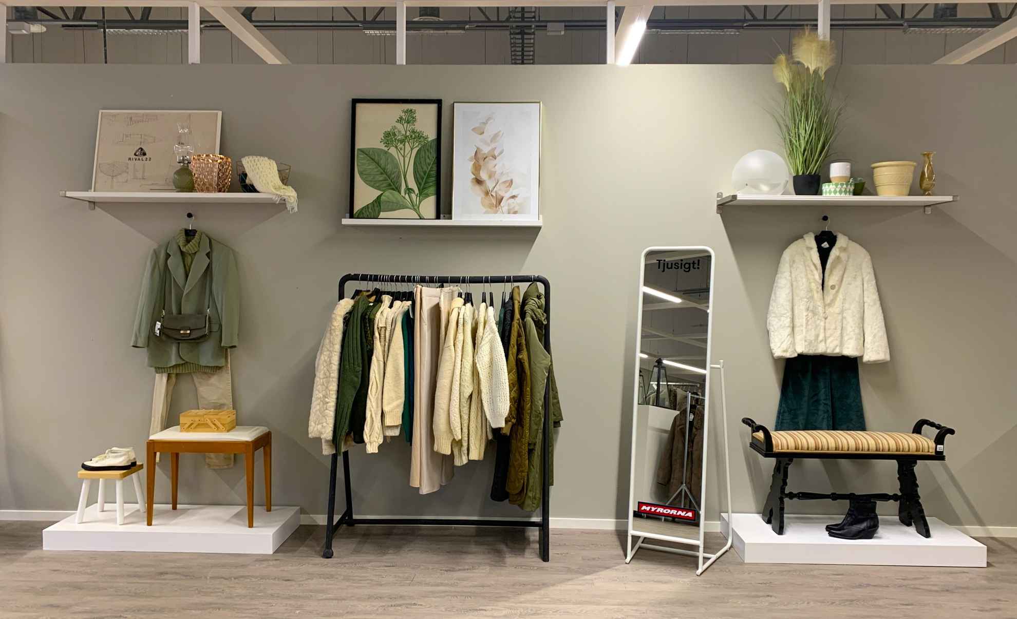 Tweedehands kleding hangt aan een rek, planken met bloemen en afbeeldingen, schoenen uitgestald en een spiegel met de winkelnaam 'Myrorna'.