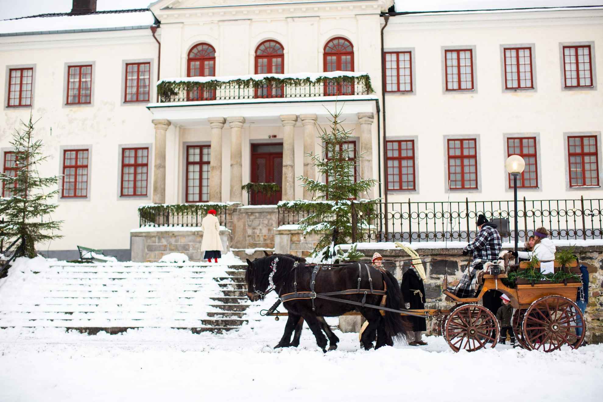 Een paard en wagen staan in de winter voor een groot wit huis. Er ligt sneeuw op de grond, een paar mensen staan naast de koets en een paar zitten erop.