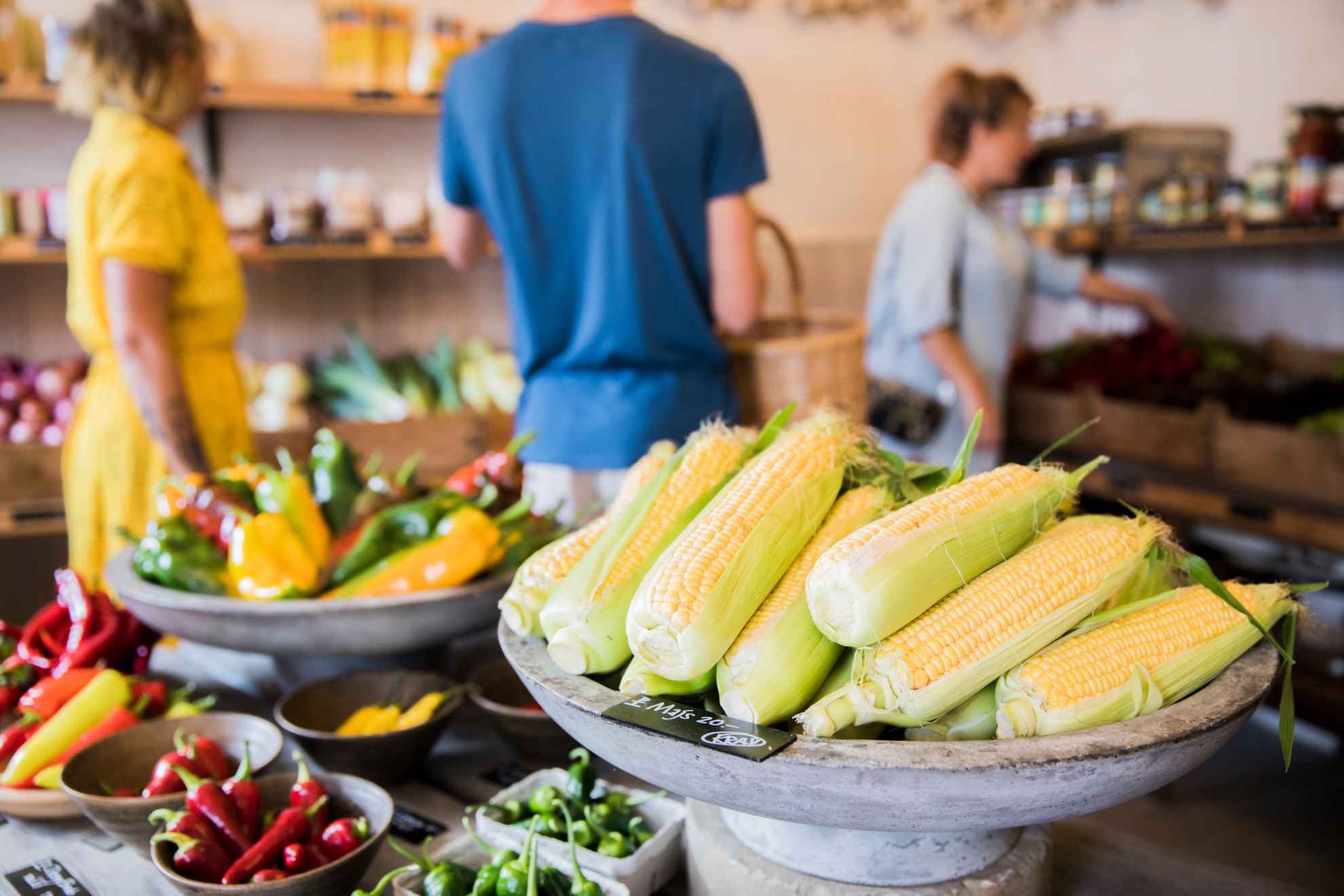 Close-up van maïskolf en een verscheidenheid aan peper in een boerderijwinkel. Op de achtergrond zijn drie personen wazig te zien.