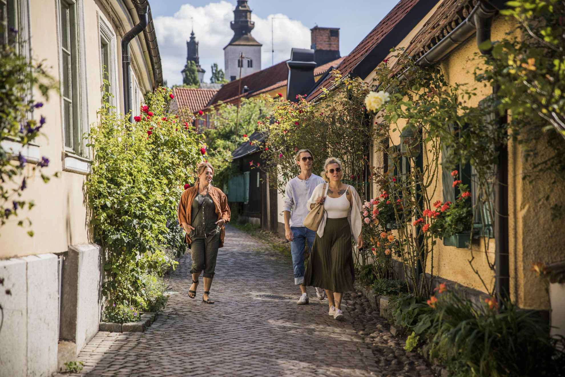 Een man en twee vrouwen slenteren door een kleine geplaveide straat in Visby. Bloemen groeien voor de oude huizen.