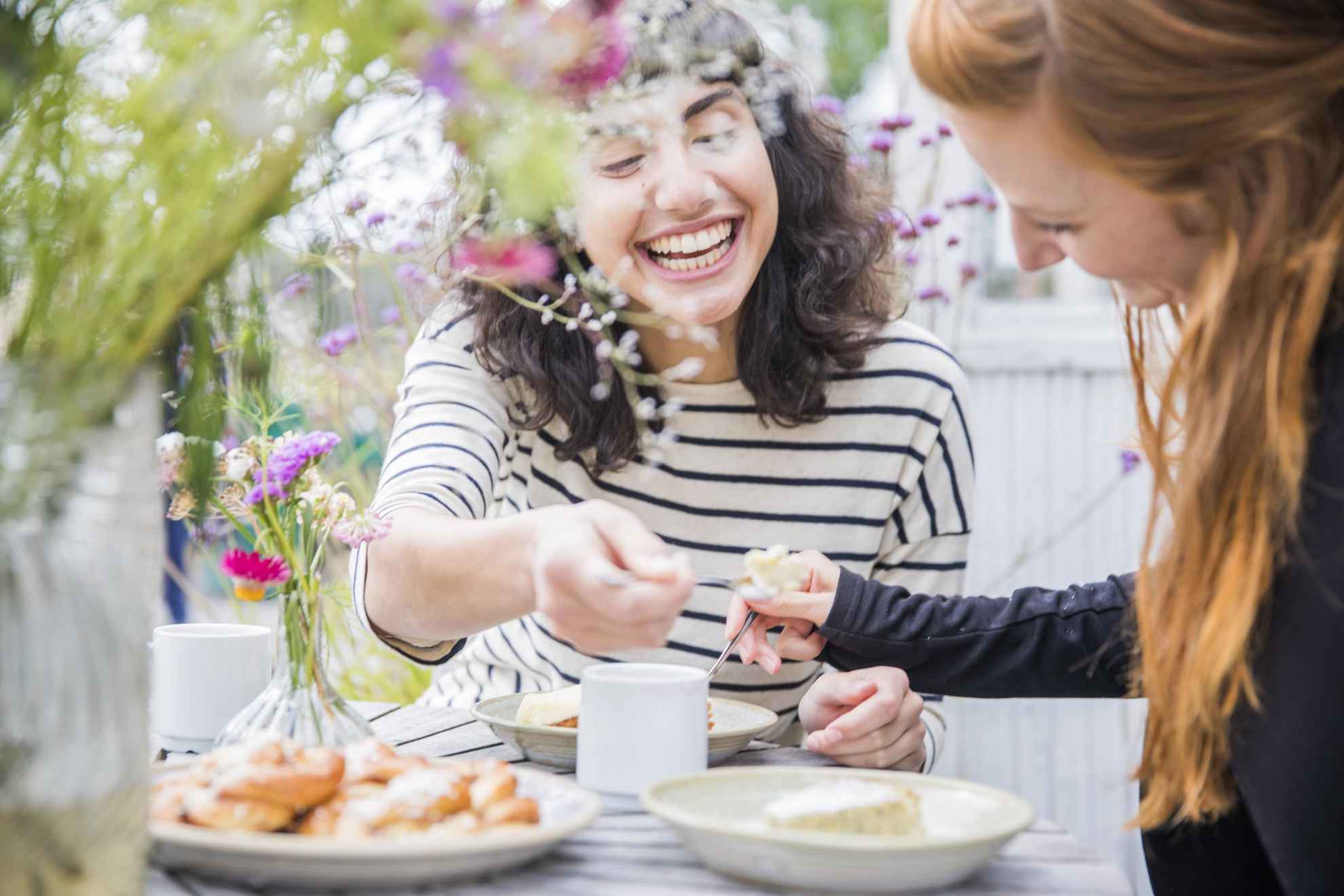 Twee vrouwen hebben een fika in de buitenlucht. Ze drinken koffie en delen taart met elkaar. Op tafel staan een bord met broodjes en twee vazen met bloemen.