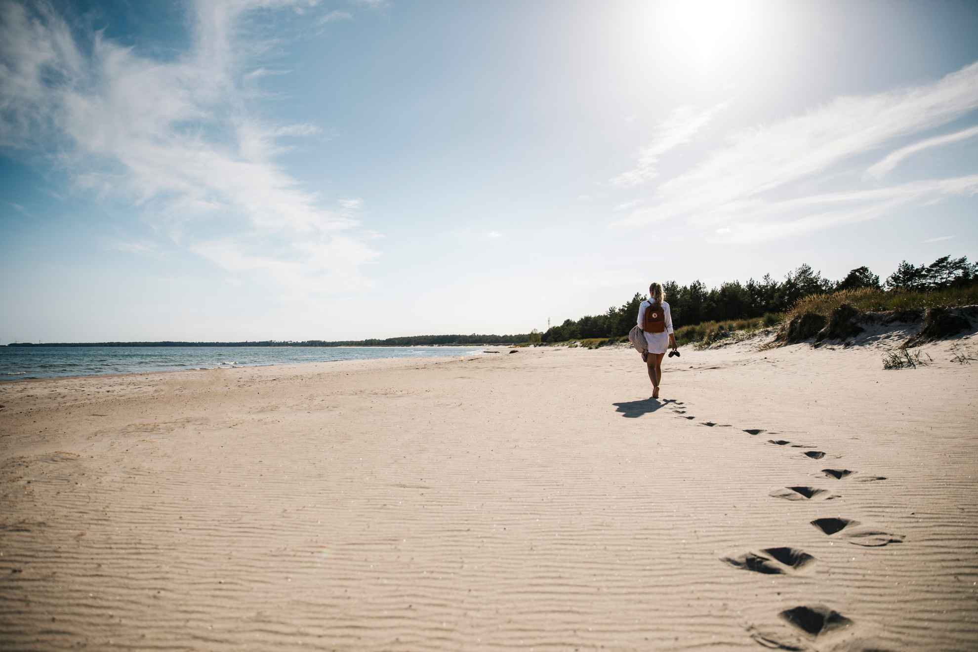 Vrouw loopt op een wit strand op Öland met een rugzak op haar rug en schoenen in haar hand.