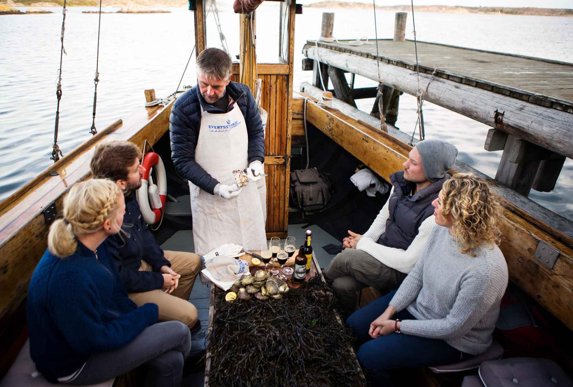 Een man gekleed in een schort toont vier mensen hoe ze oesters moeten openen op een boot. Talrijke oesters op een tafel bij hen, en een glas met drank.