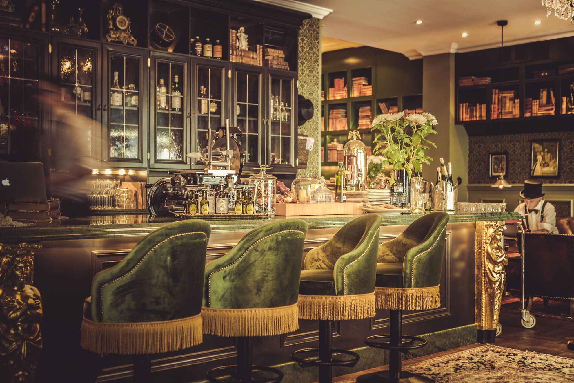 De bar van hotel Pigalle, met groenfluwelen stoelen, houten planken en gouden details. Iemand beweegt achter de bar.