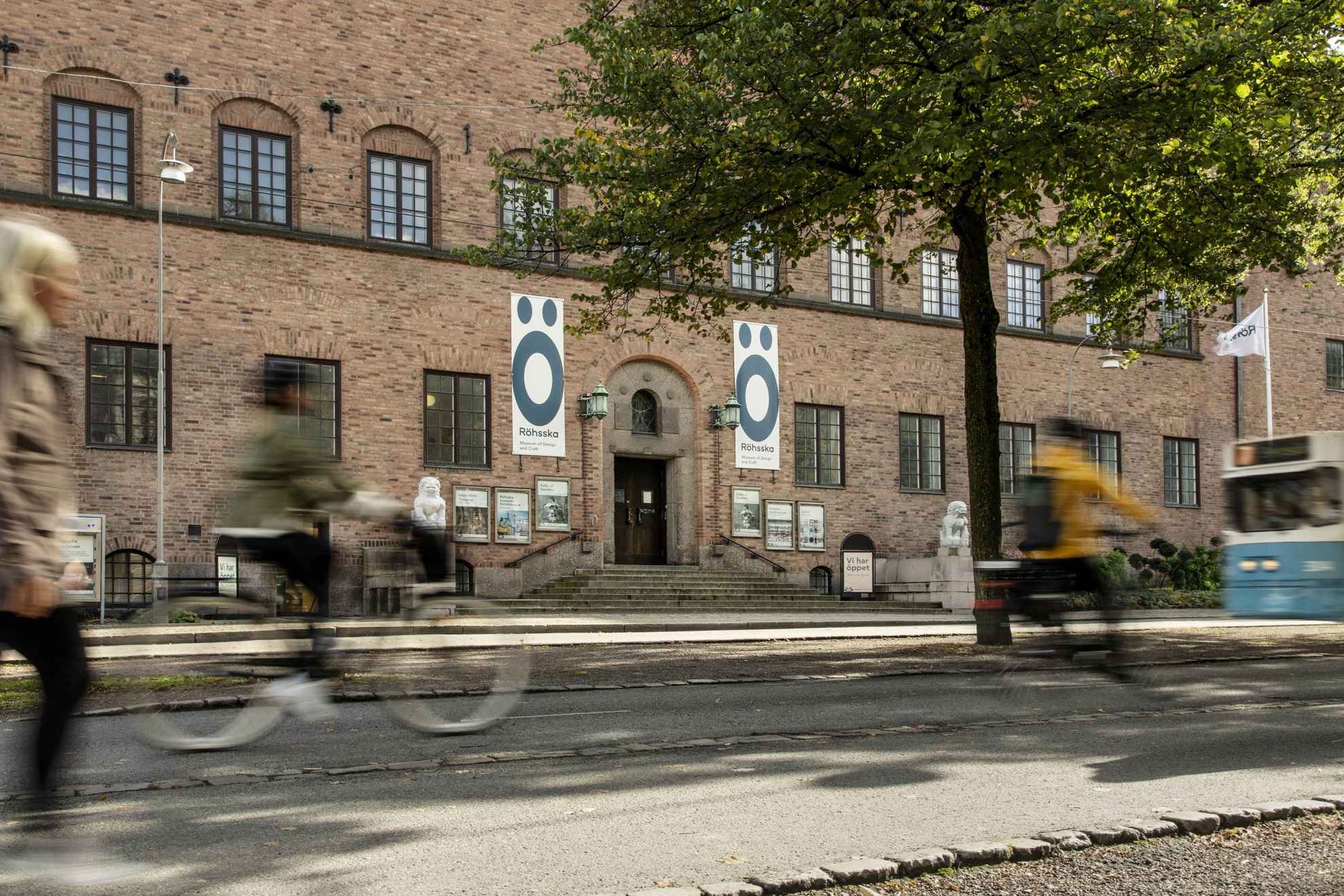 Twee fietsende en een wandelende persoon worden wazig gezien langs de ingang en de gevel van het Röhsska-museum in Göteborg. Het gebouw is gemaakt van bakstenen en aan elke kant van de ingang staan witte stenen beelden met figuren die een beetje op leeuwen lijken.