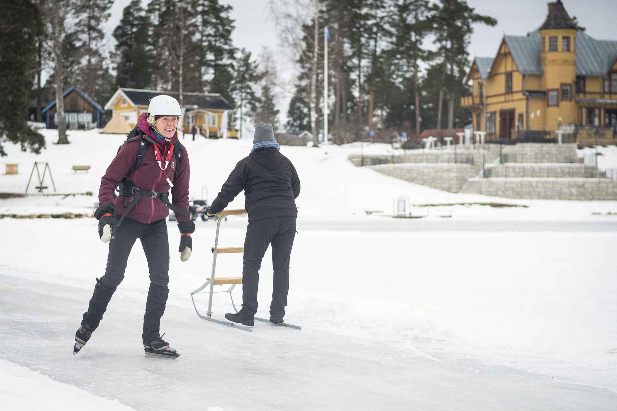 Een vrouw schaatst lange afstanden terwijl een andere persoon achter haar een slee op het ijs duwt.