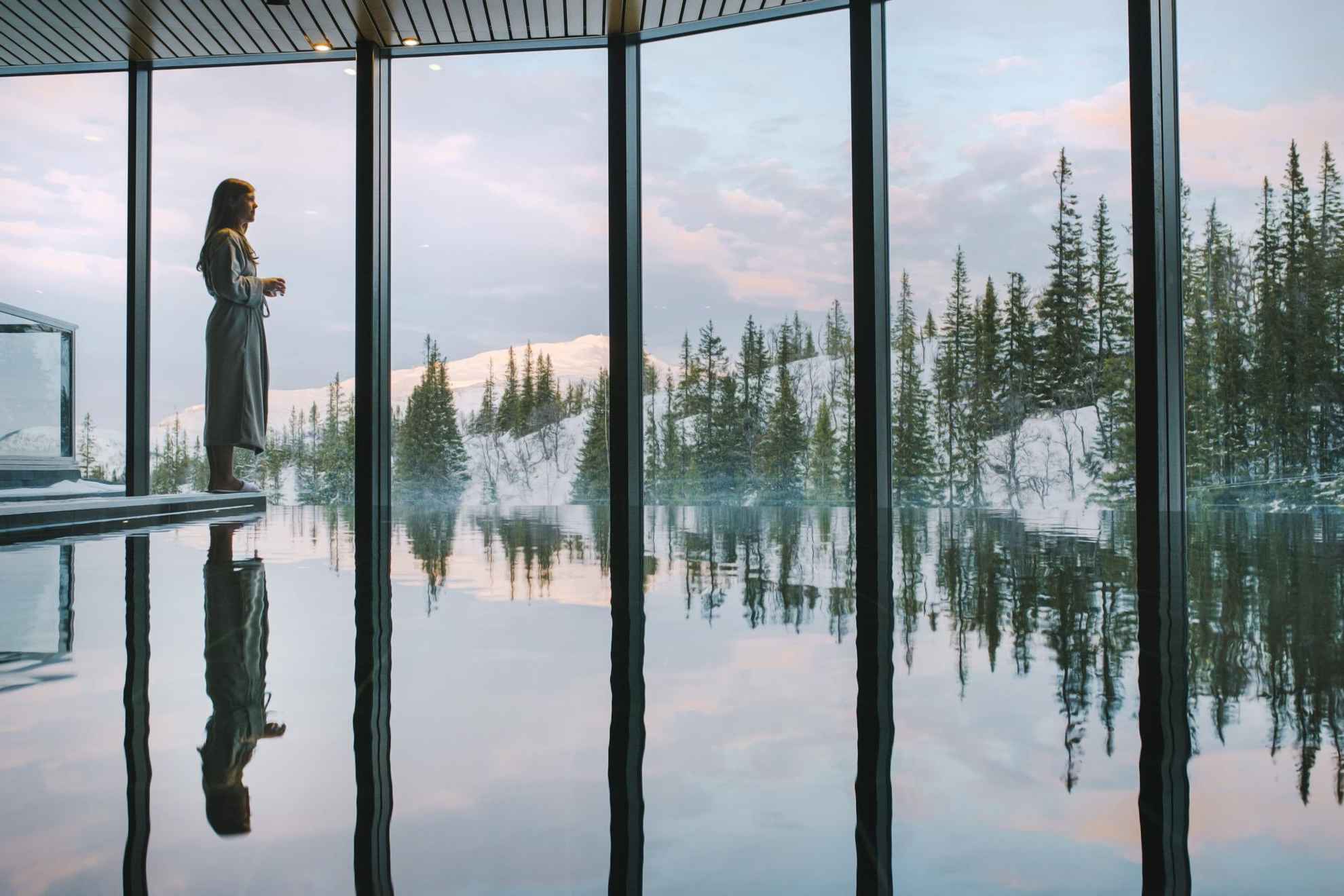 Een vrouw die aan het zwembad staat in een spa met grote ramen die uitkijken op de bergnatuur.