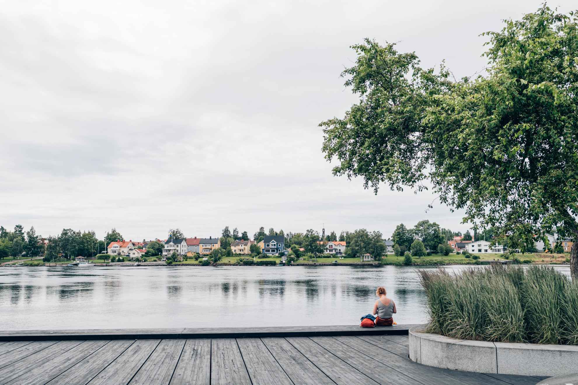 Een vrouw zit op de promenade langs een rivier. Aan de overkant van de rivier zie je mooie villa's in een groene omgeving.