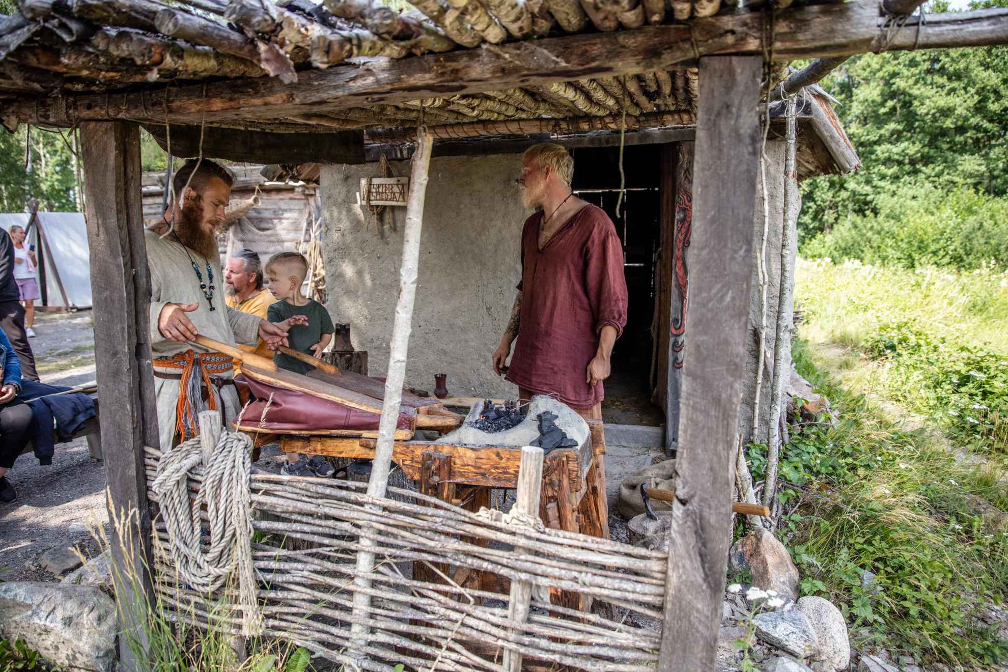 Twee kinderen kijken toe hoe twee mannen, gekleed in historisch typische vikingkleding, aan het werk zijn bij een haard.