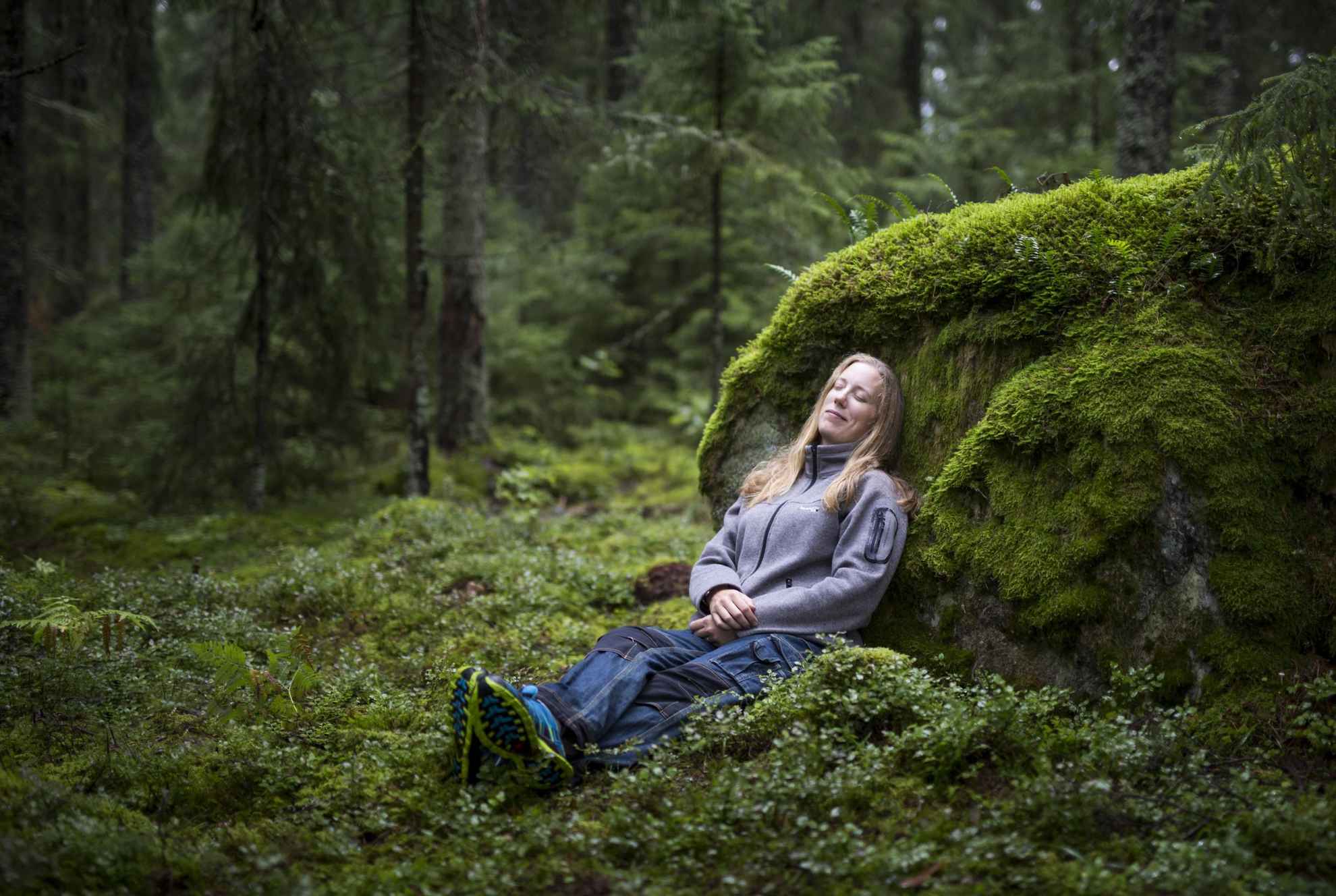 Een vrouw zit op de grond in een bos en rust tegen een rotsblok bedekt met mos.