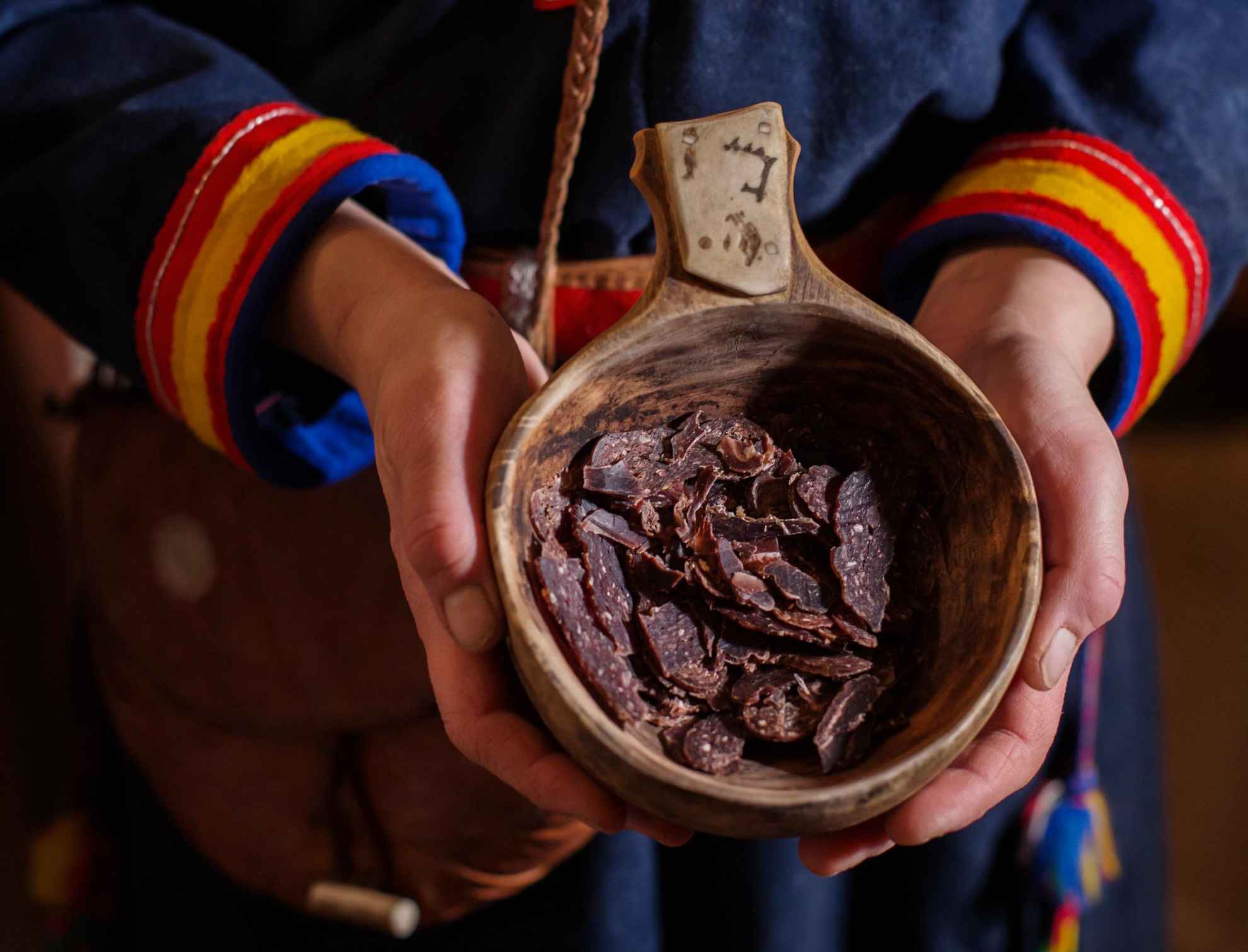 Een persoon gekleed in Sámi-kleding met een houten beker gevuld met stukjes gedroogd rendiervlees.
