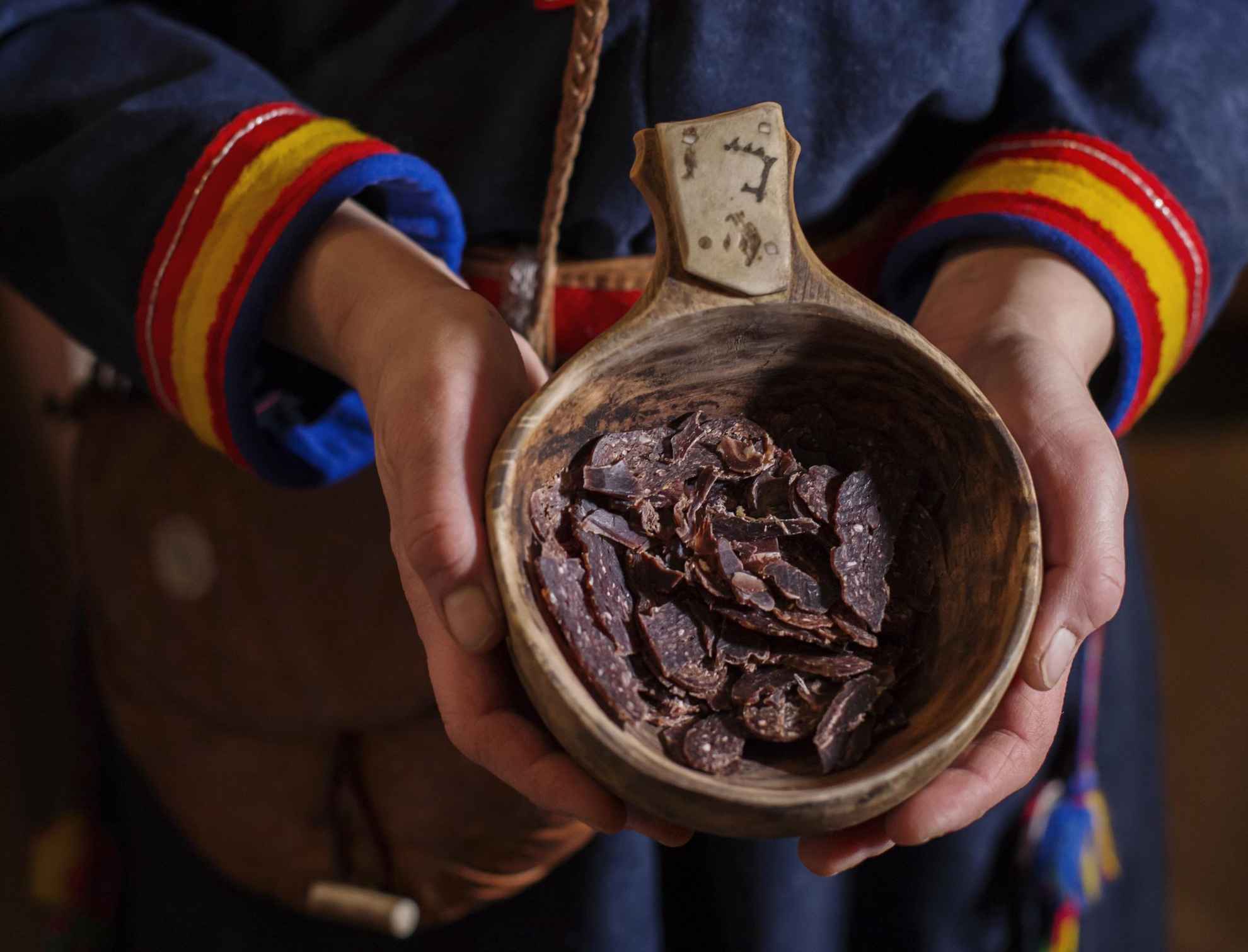 Een persoon gekleed in Sámi-kleding die een houten beker vasthoudt gevuld met stukjes gedroogd rendiervlees.