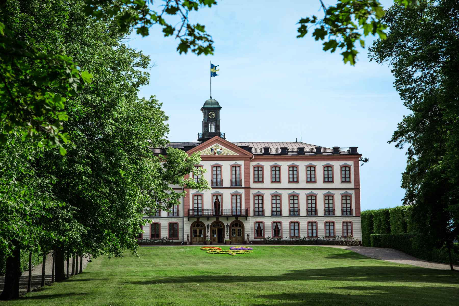 De buitenkant van Kasteel Tullgarn met witte muren en grote ramen omringd met rode details. Bomen en een grasveld omringen het gebouw.