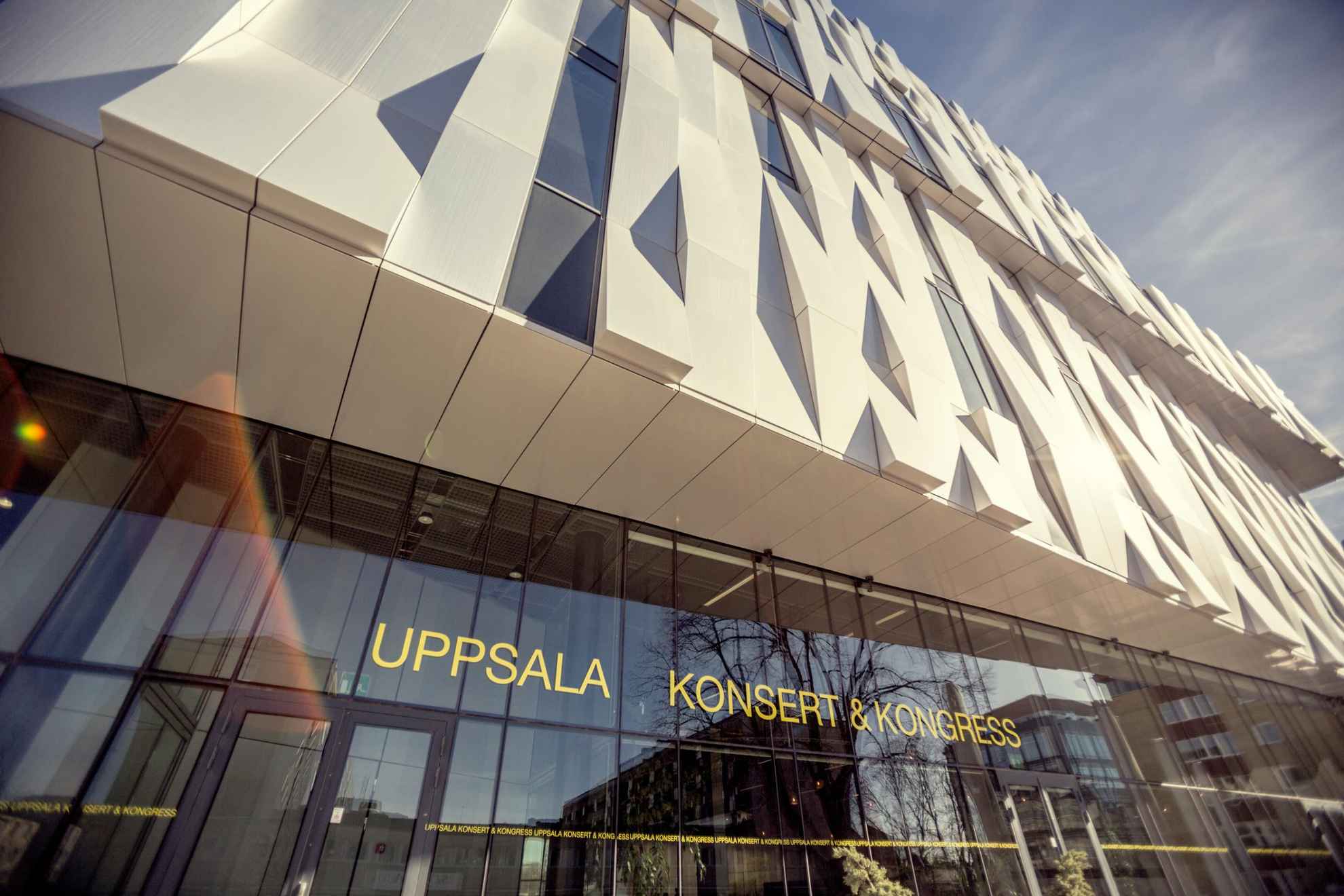 Een close beeld op een onderscheidend gebouw, met een kristalachtige gevel bestaande uit reflecterende metalen platen. Bij de raammuur op de benedenverdieping staat een bord met de tekst Uppsala Konsert & Kongress.