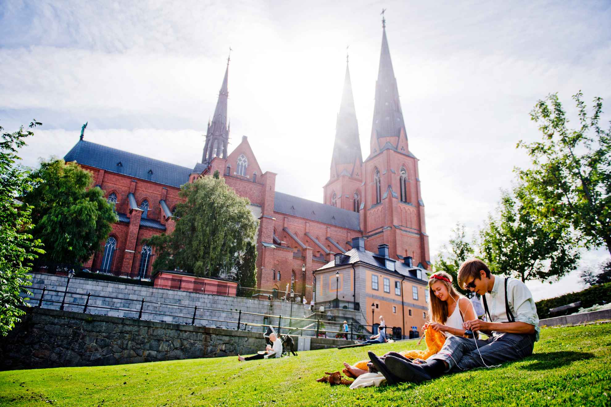 Zonnig uitzicht over de kathedraal van Uppsala met mensen op de voorgrond zittend op het groene gras.