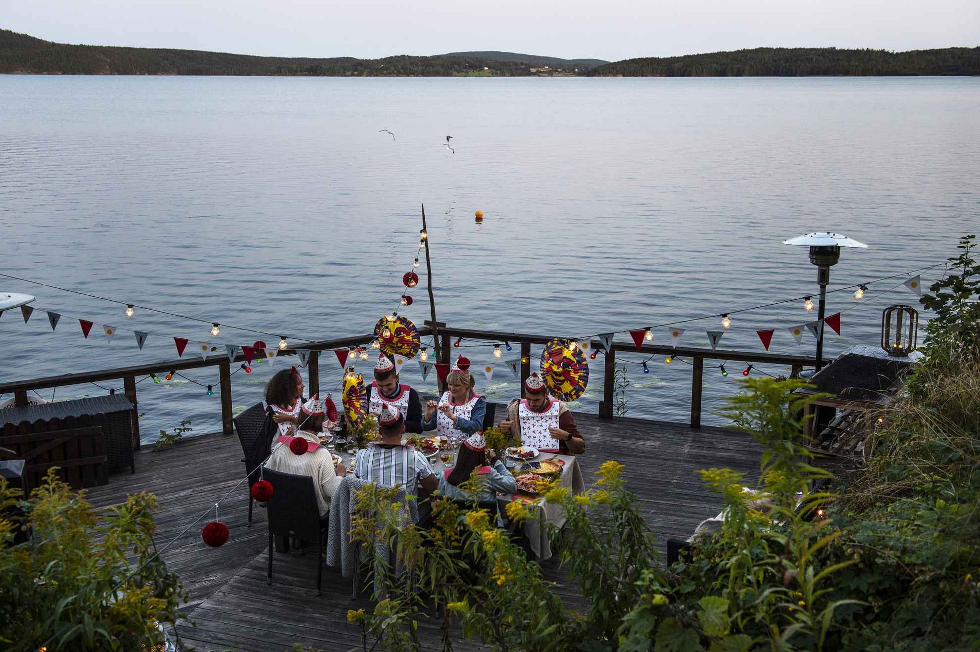 Rivierkreeftenfeestje op een steiger aan zee. Zeven mensen rond een tafel gekleed in papieren hoeden en slabbetjes eten rivierkreeft. Boven de tafel hangen papieren lantaarns, lampjes en versieringen.