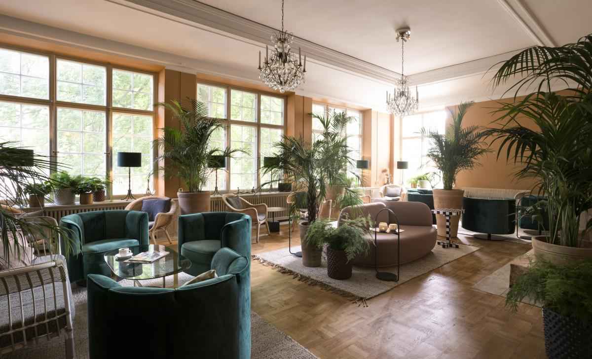 Een zithoek met banken en fauteuils. De kamer is versierd met planten en twee kroonluchters die aan het plafond hangen.
