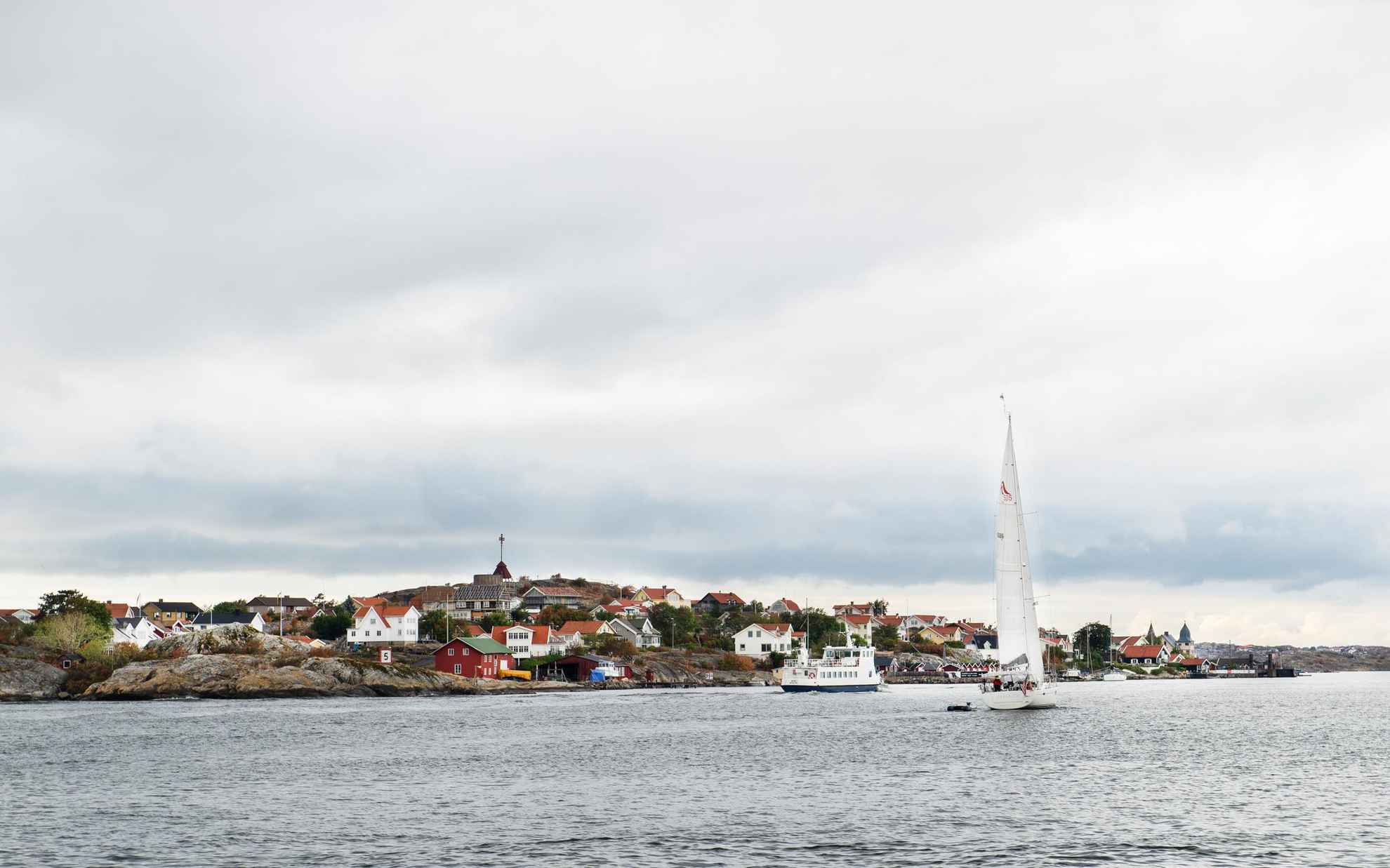 Een kleine veerboot en zeilboot in de zee, langs een eiland met veel huizen buiten Göteborg.