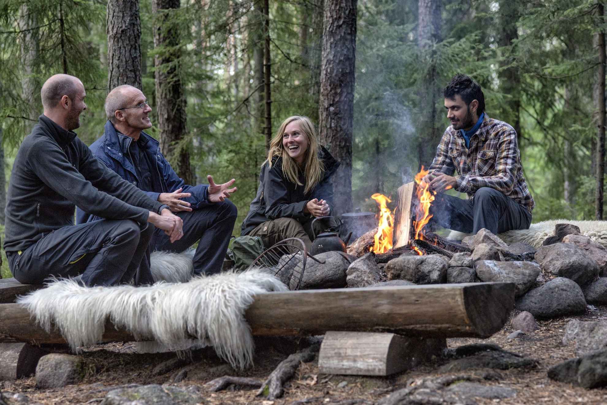 Vier mensen zitten lachend rond een kampvuur in een bos.