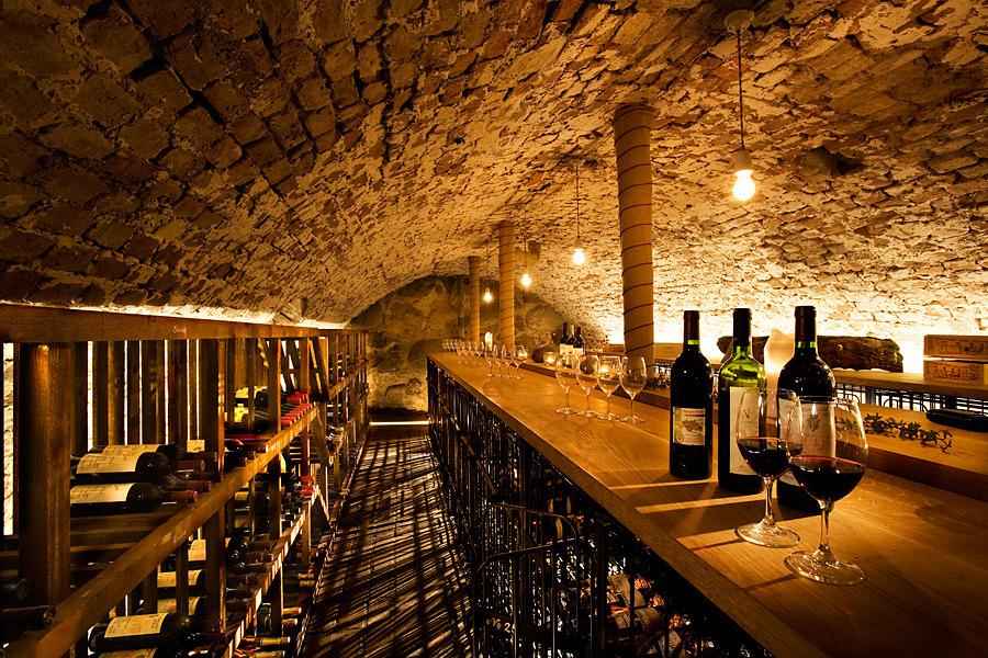 De wijnkelder van Ulfsunda Slott heeft een rond bakstenen plafond. Flessen en wijnglazen op een tafel in het midden van de kamer. En flessen op planken aan de linkerkant.