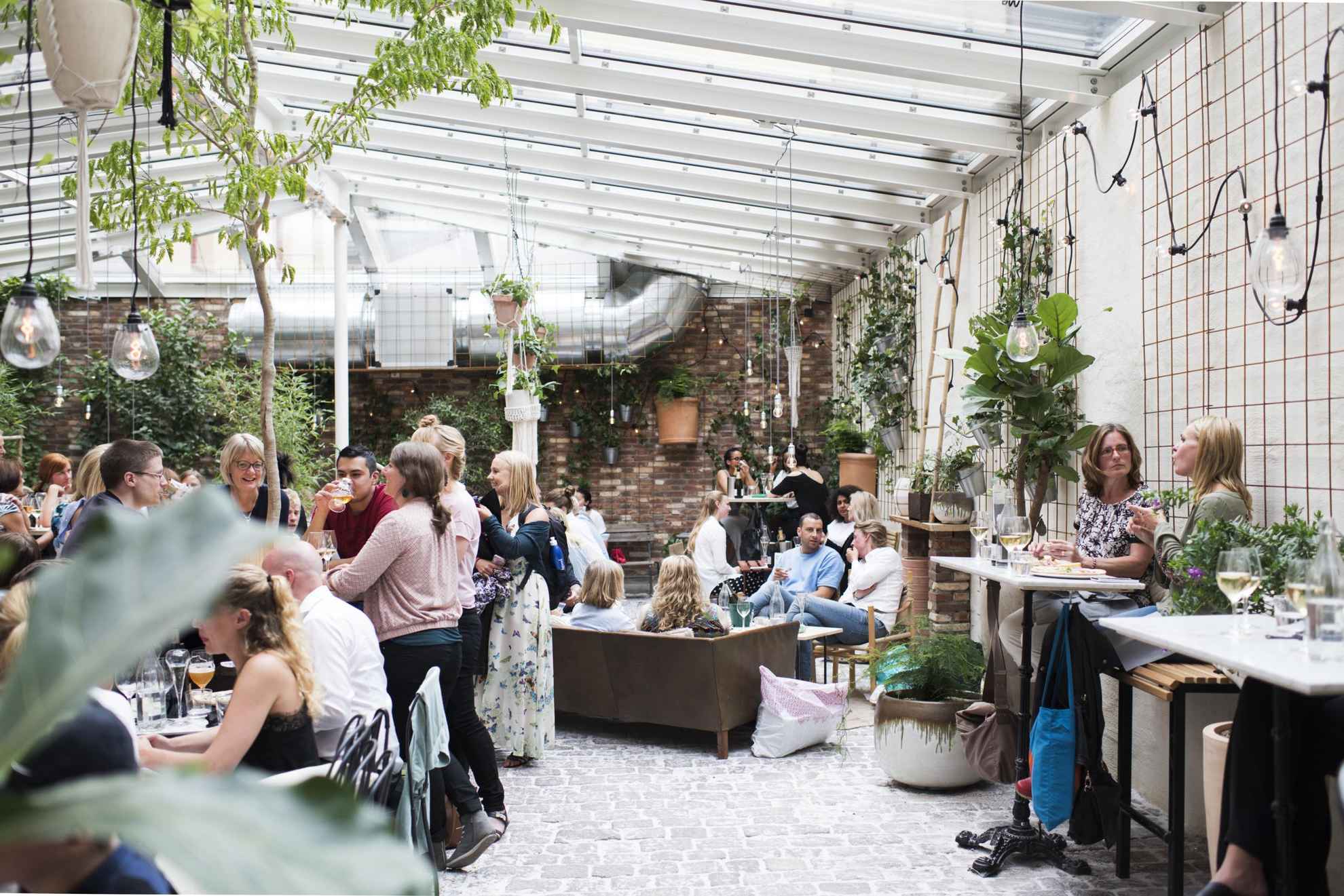 Mensen genieten van drankjes en eten in een druk café Magasinet in Göteborg. Het interieur is een stenen vloer, bakstenen muren, een glazen plafond en de ruimte staat vol met groene planten.
