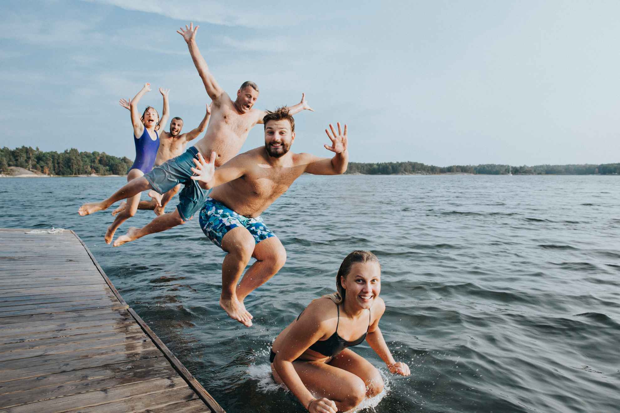 Vijf jonge mensen in badkleding springen vanaf een houten pier in een meer.
