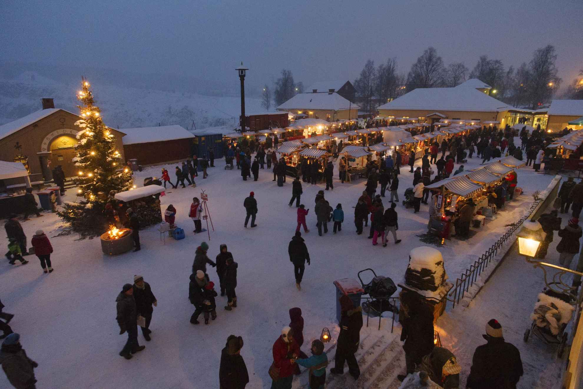Een kerstmarkt in de sneeuw, met veel kraampjes, mensen en een grote kerstboom bij een klein kampvuurtje.