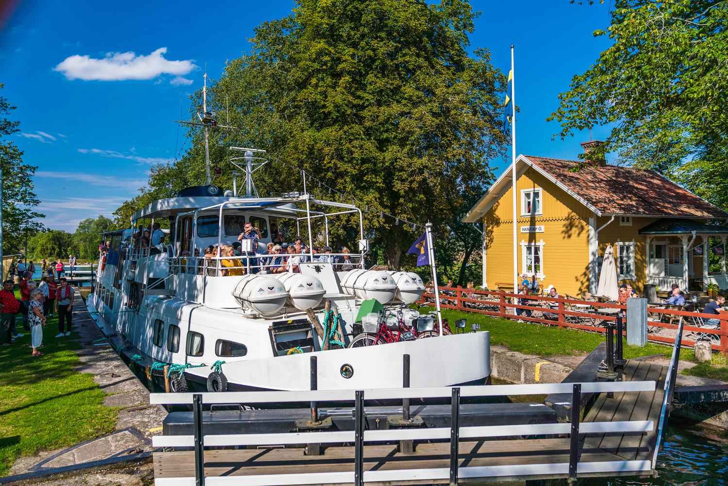 Een grote witte boot passeert het gele houten huis van hostel en café Hajstorp.