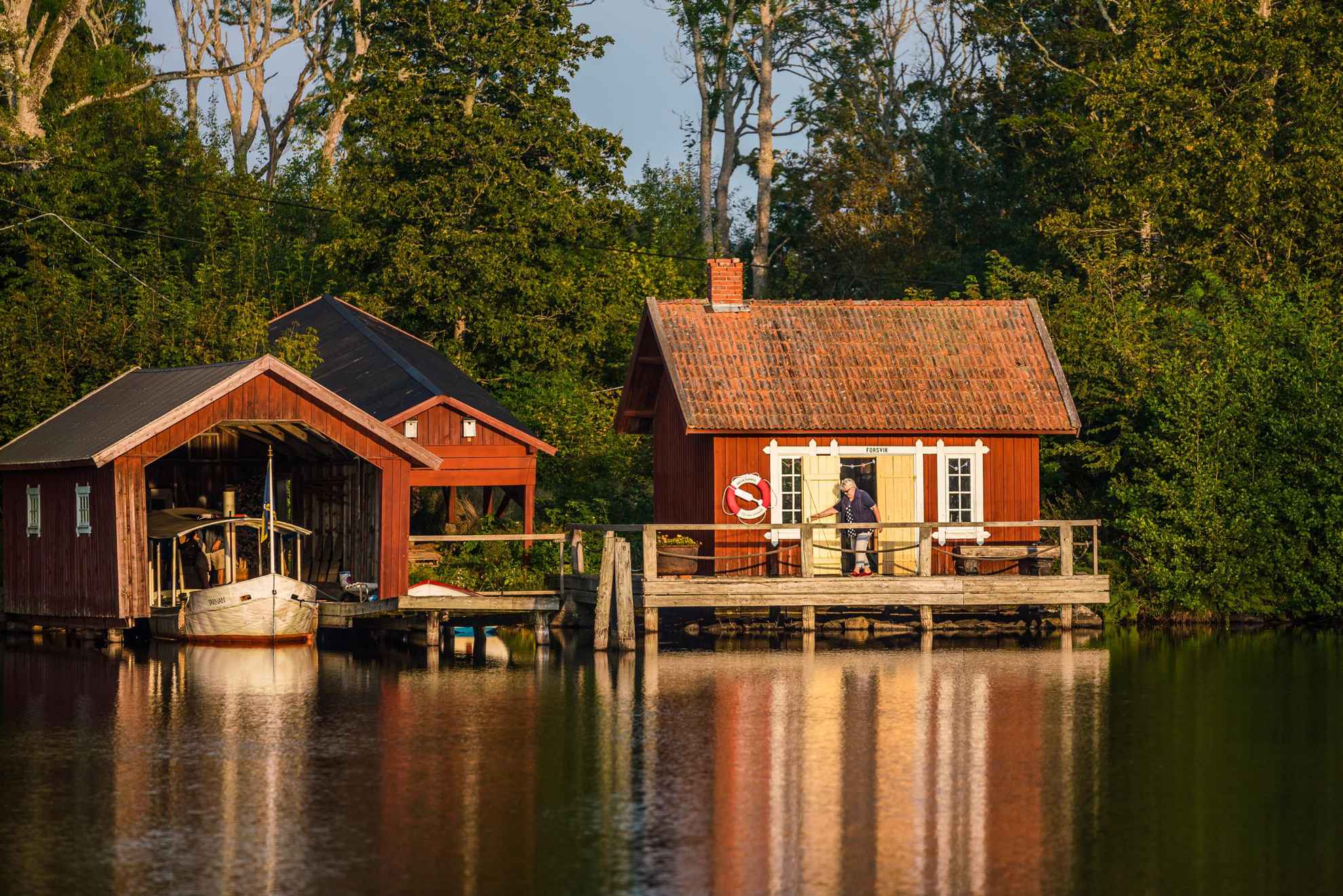 Een vrouw verlaat een rood houten huis dat aan het water ligt. Naast het huis staat een botenhuis.