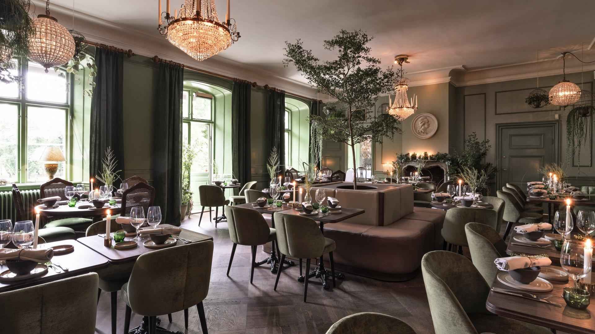 Een grote eetkamer ingericht met houten tafels, groene stoelen en groene muren. Aan het plafond hangen kroonluchters.