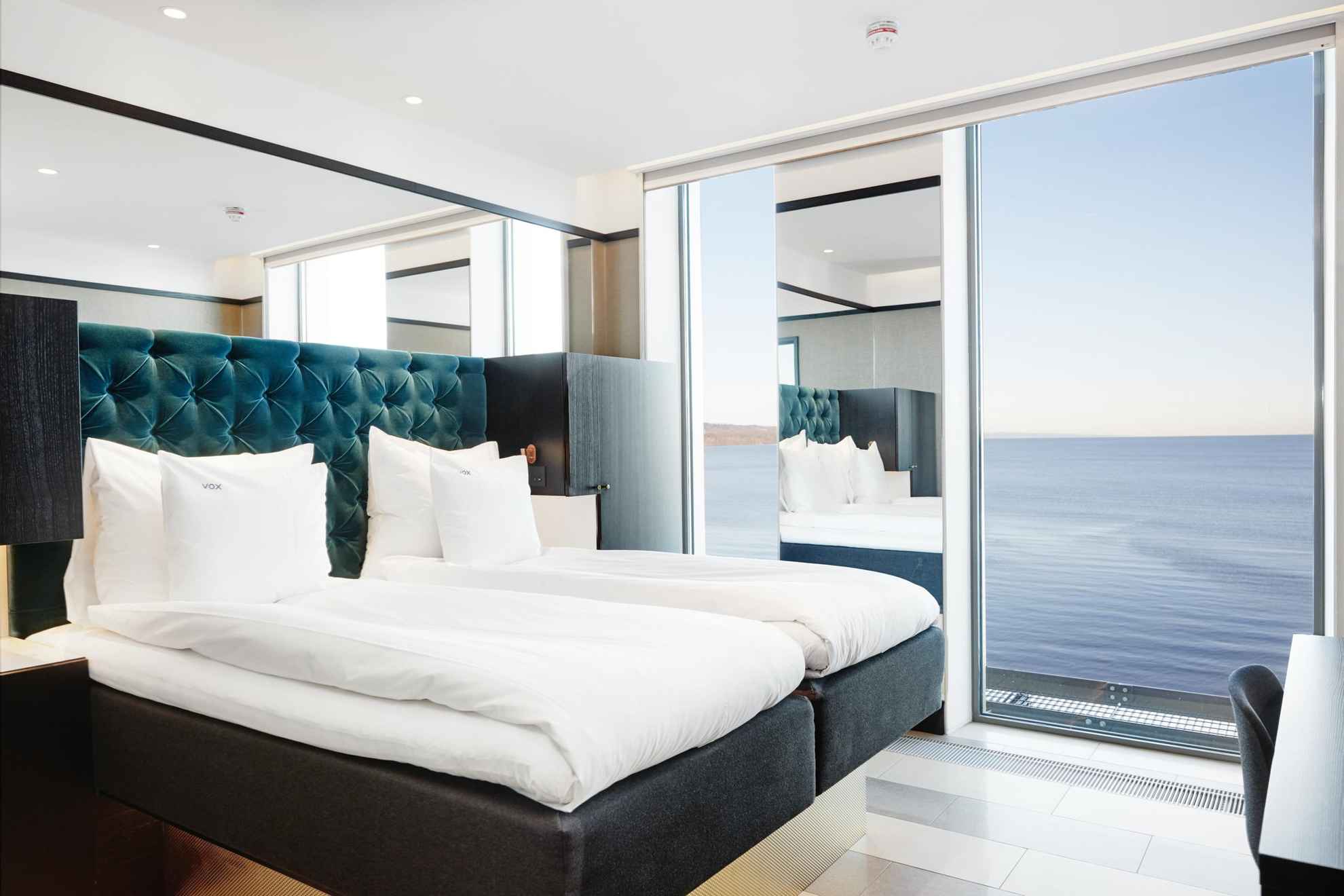 Een hotelkamer met een tweepersoonsbed met witte lakens. De kamer heeft grote ramen met uitzicht op het meer Vättern.