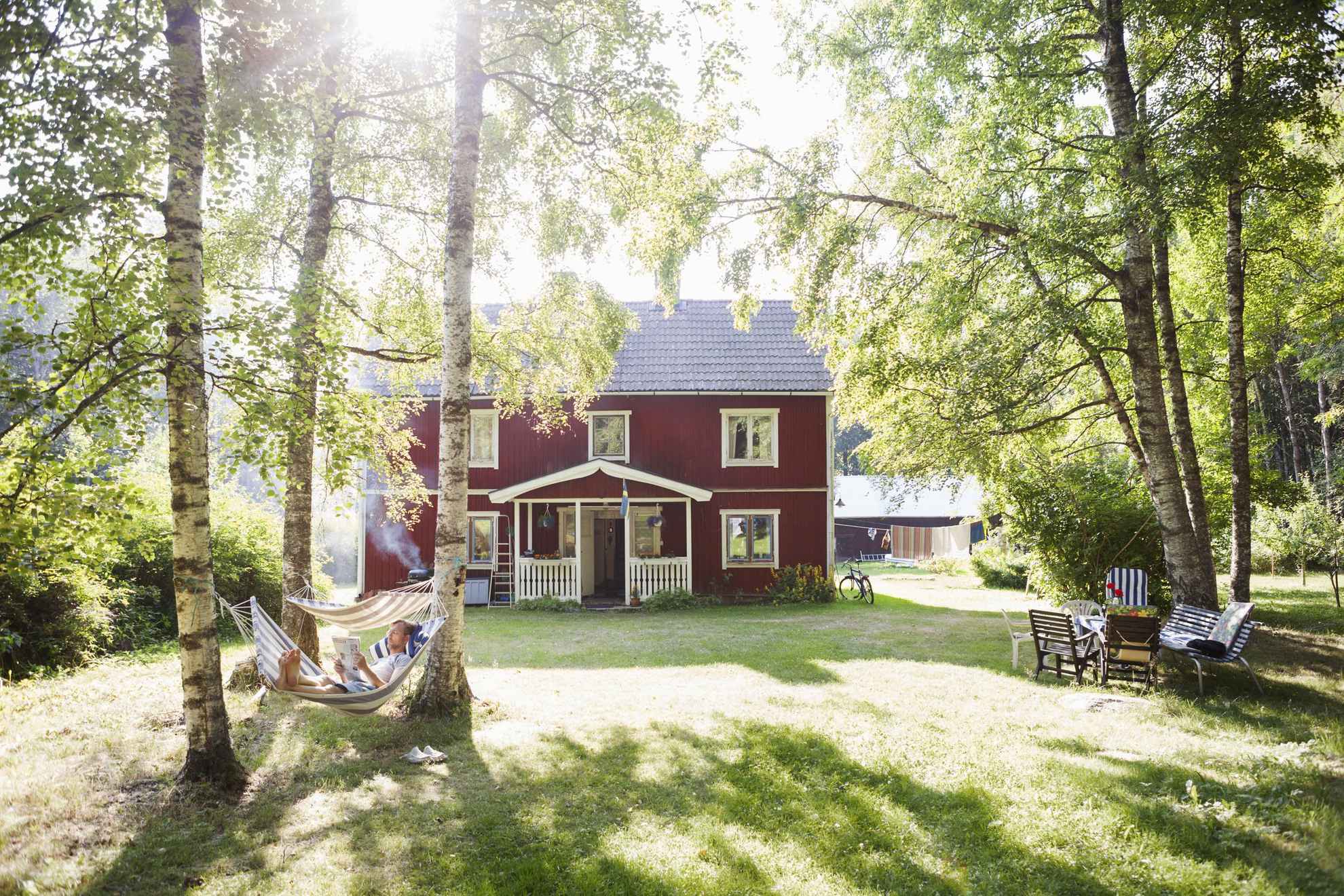 Een tuin voor een traditioneel rood Zweeds huis, met groen, bomen, hangmatten en tuinmeubelen.