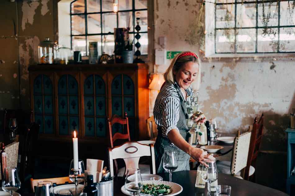 Een vrouw zet flessen op tafel in een restaurant. De tafel is gedekt met borden, wijnglazen en een schaal met salade. Op de achtergrond staat een oude kast.