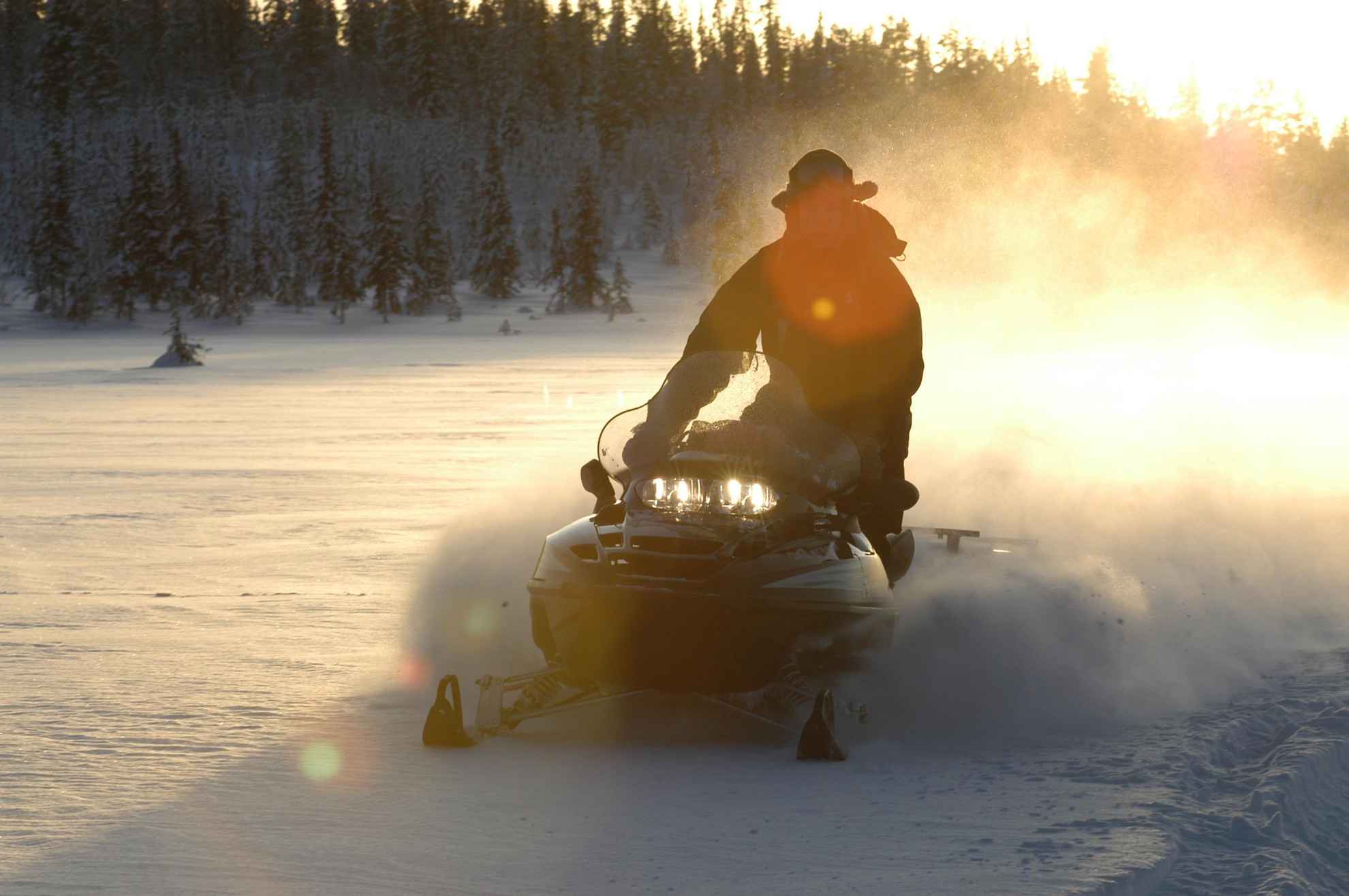 Een persoon op een sneeuwscooter in een besneeuwd landschap bij laagstaande zon.