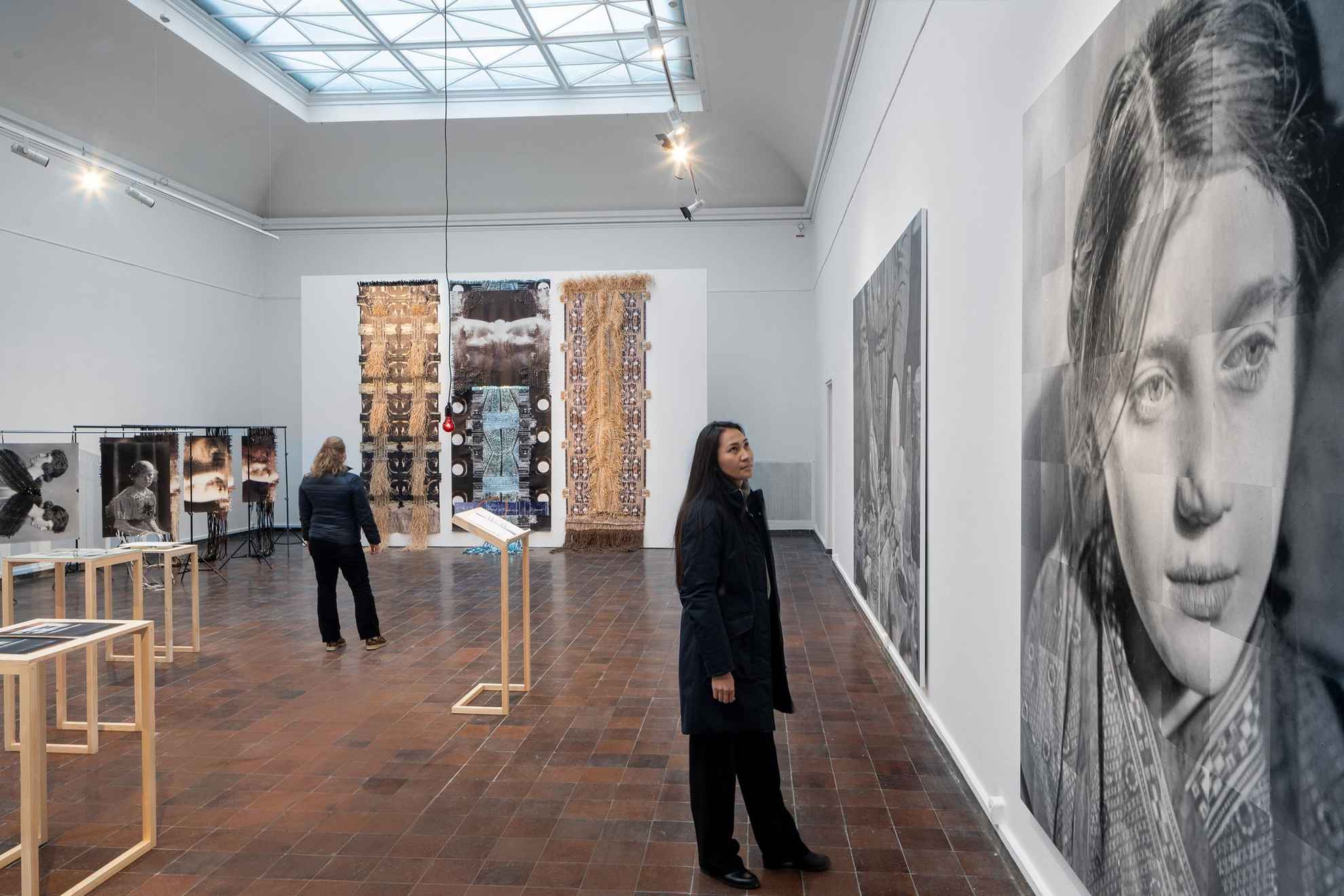 Een installatie op een tentoonstelling. Een vrouw kijkt naar een grote zwart-wit afbeelding van een vrouw en op de achtergrond kijkt een vrouw naar een groot kunstwerk met foto's die met fragmenten aan elkaar zijn gevlochten tot nieuwe structuren.