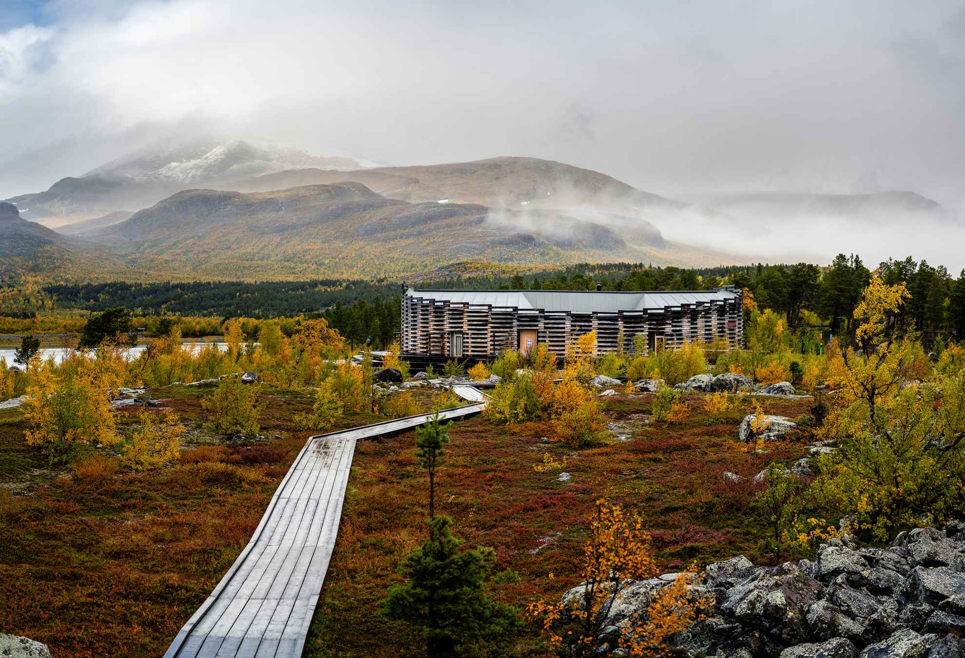 Een houten loopbrug naar het Naturum Laponia-gebouw. Achter het gebouw zijn bergen die licht bedekt zijn met mist.