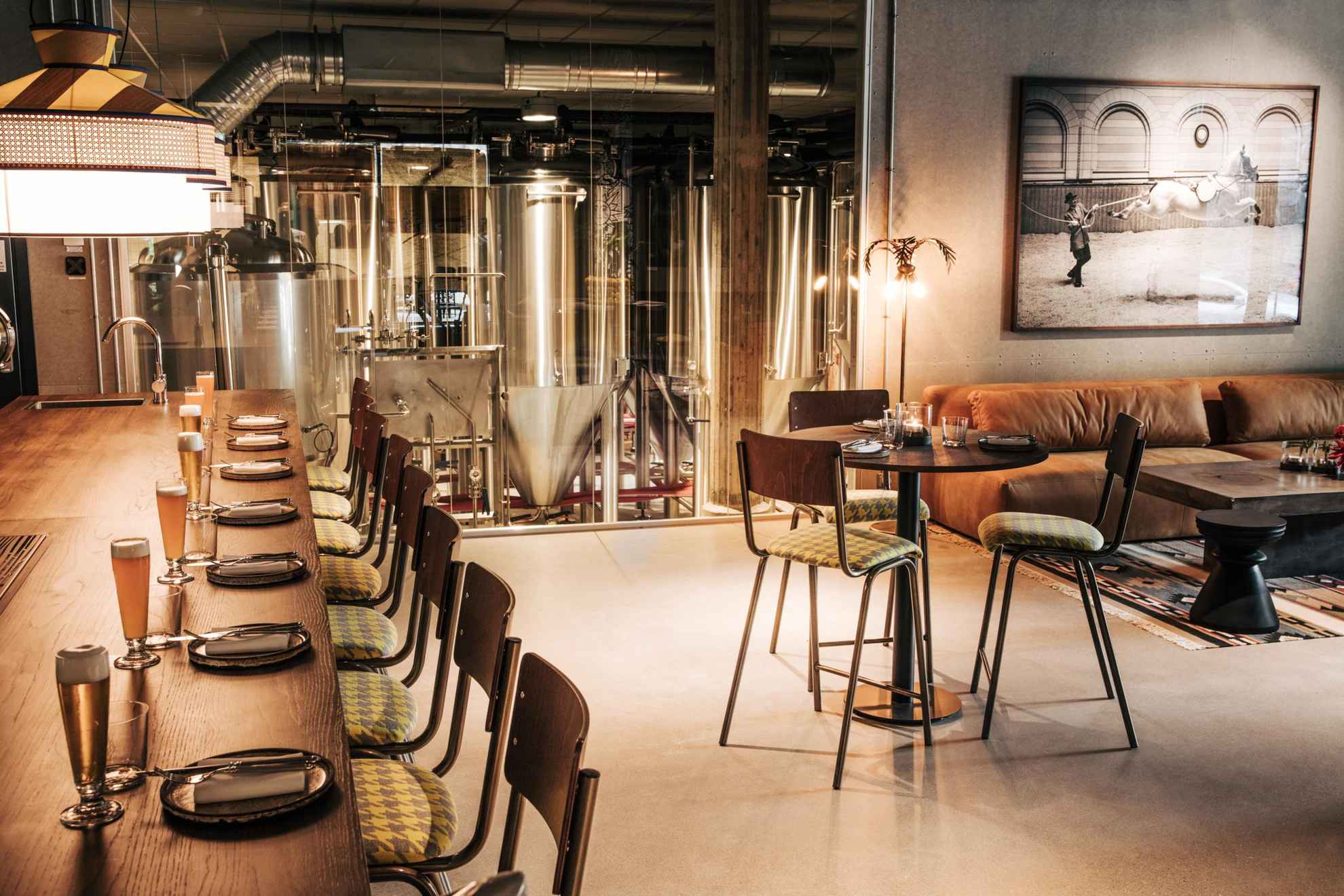 In een restaurant staat links de bar met glazen bier, rechts een gedekte tafel met drie stoelen en een bank. Op de achtergrond achter een glazen wand zie je een brouwerij.