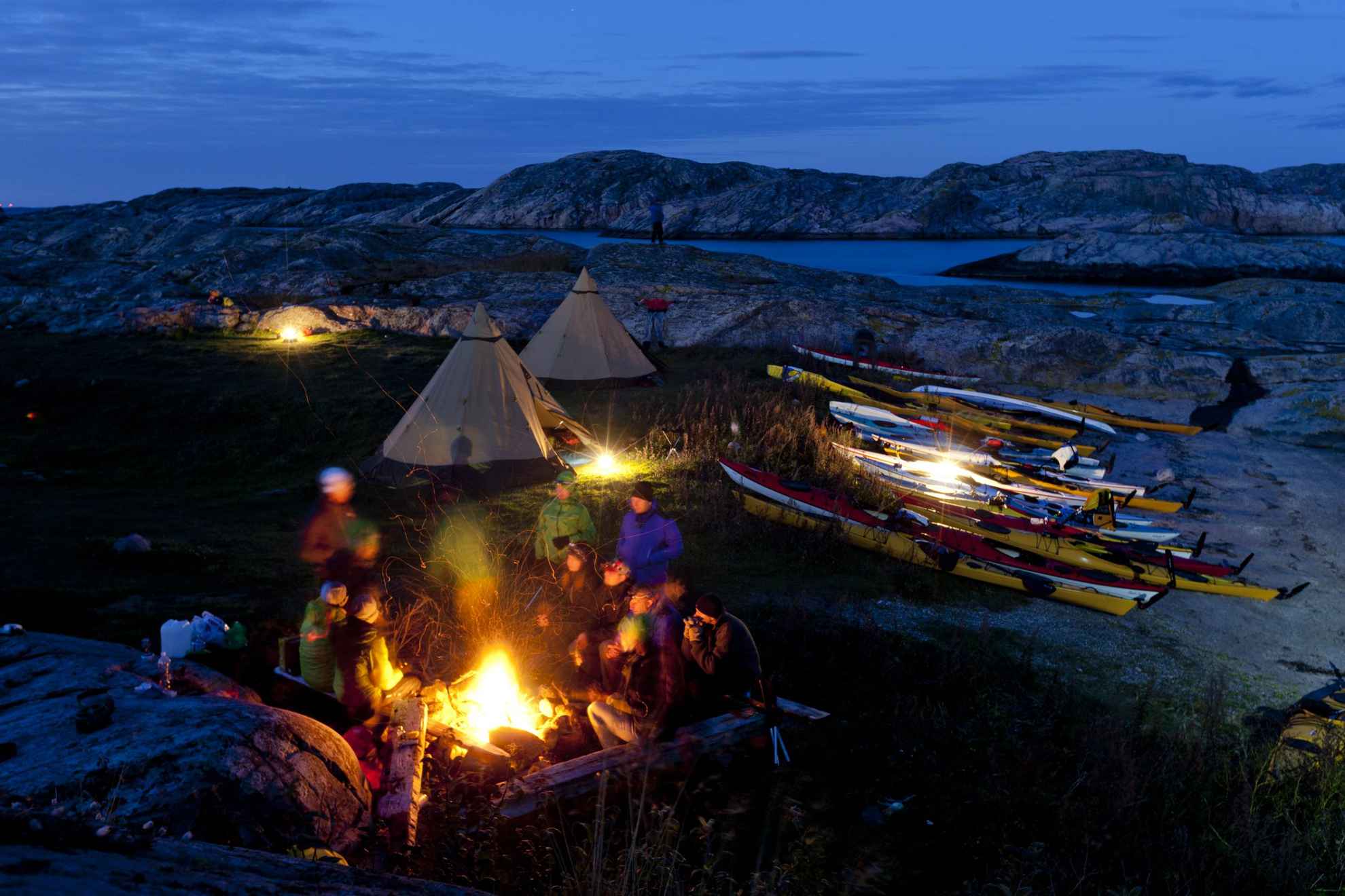 2 tenten zijn opgesteld op rotsen. Naast de tenten liggen kayaks en er brandt een open vuur. Het is na zonsondergang.