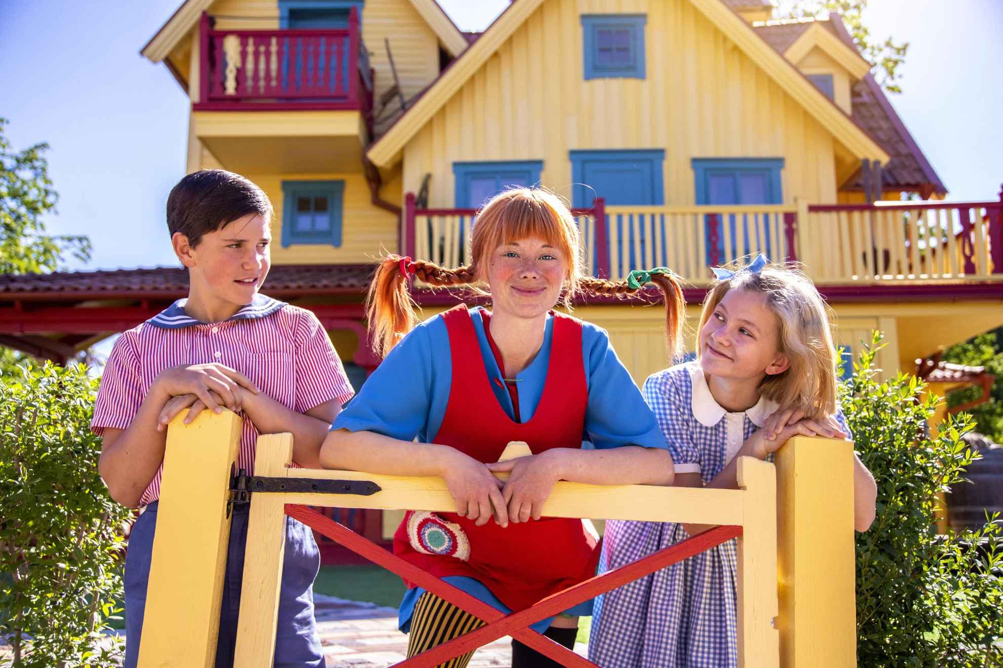 Acteurs verkleed als de personages Tommy, Pippi en Annika staan bij een gele poort voor een geel huis (Villa Villekulla).