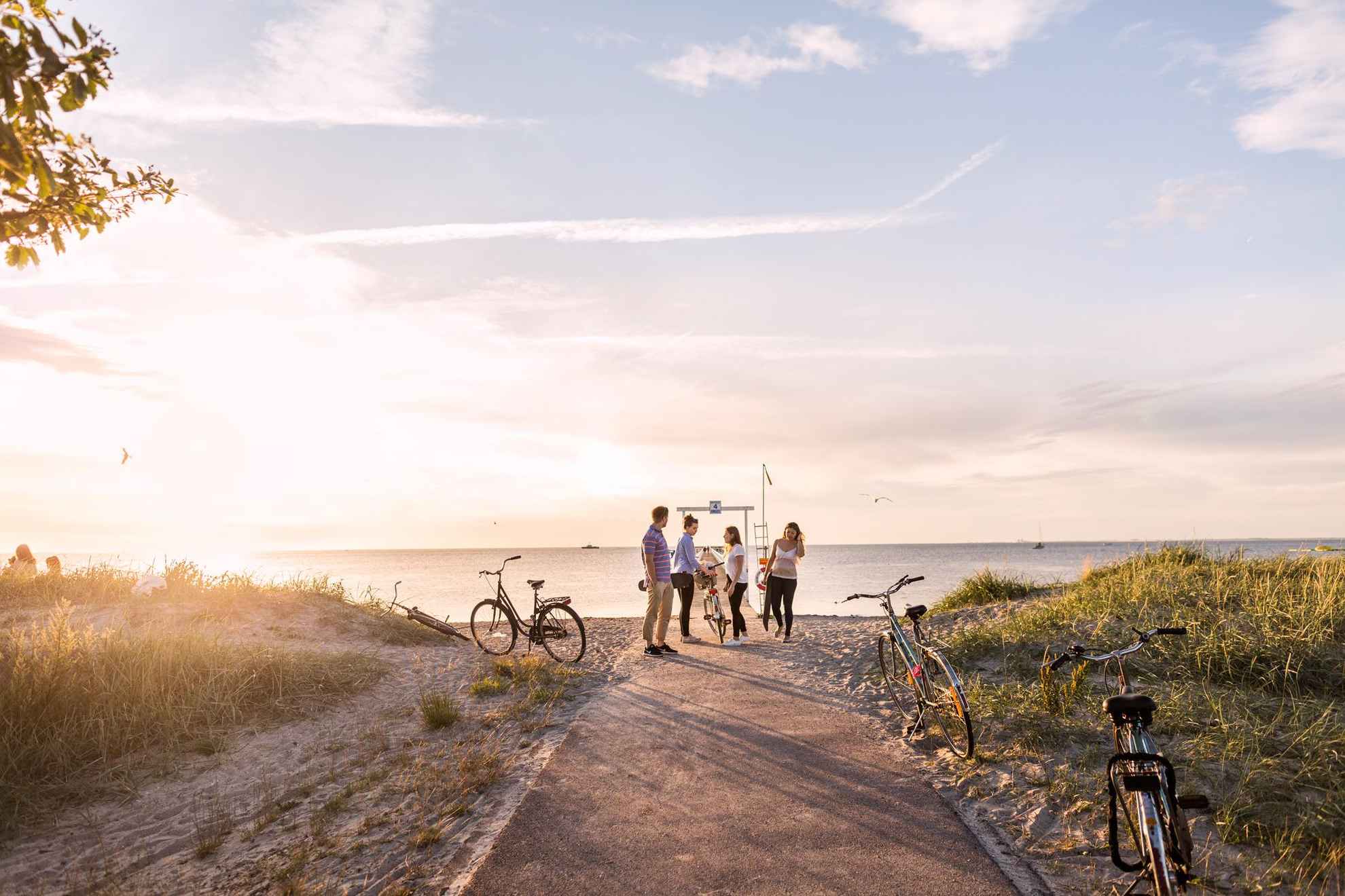 Zonsondergang op het strand van Ribersborg, een man en drie vrouwen worden gezien bij een zwemsteiger op het strand. Er staan serverfietsen geparkeerd langs het pad naar het strand.