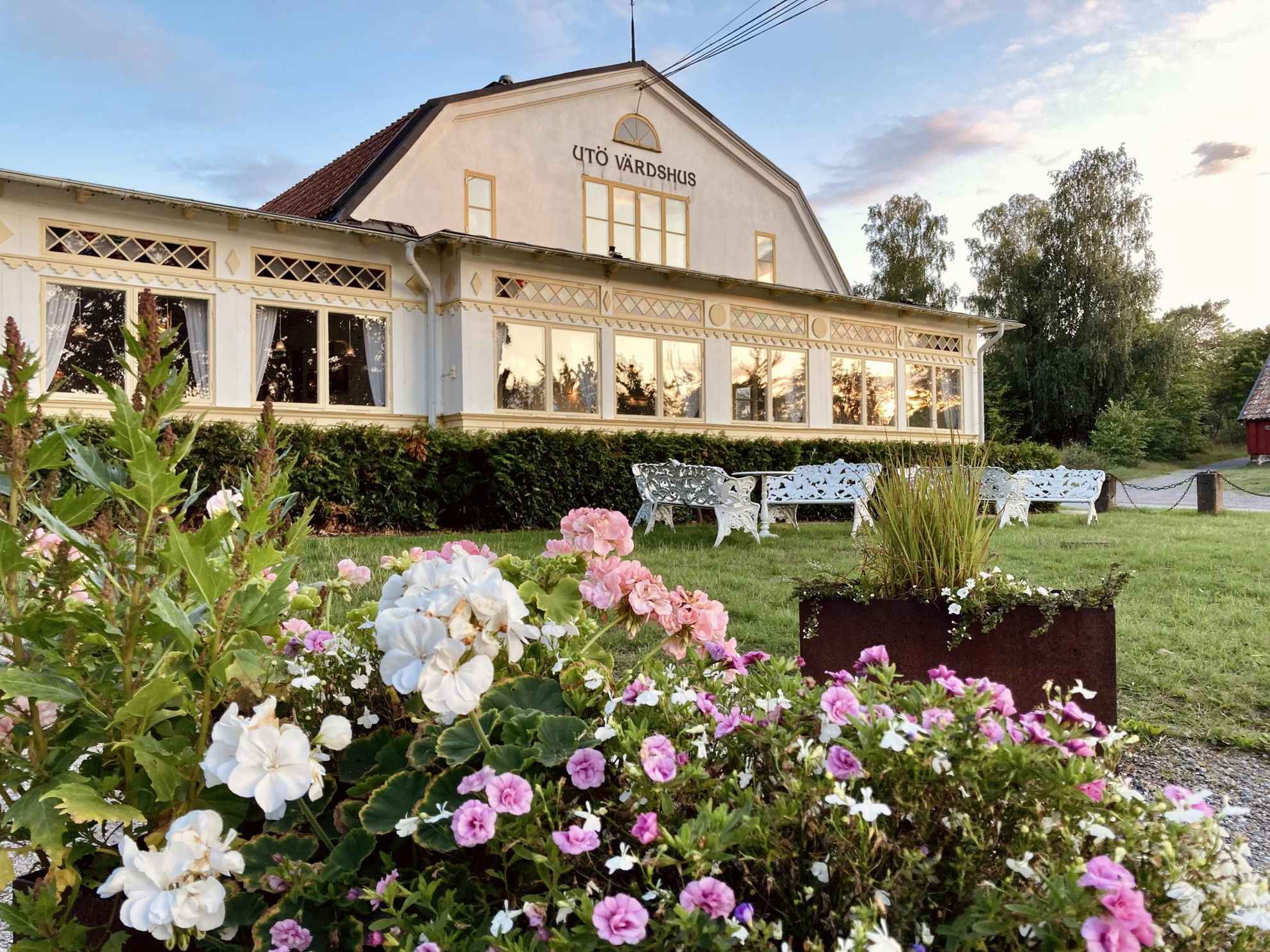 Een witte villa uit de eeuwwisseling met lichtgele uitgesneden details rond de ramen. Struiken met roze bloemen zijn zichtbaar op de voorgrond.