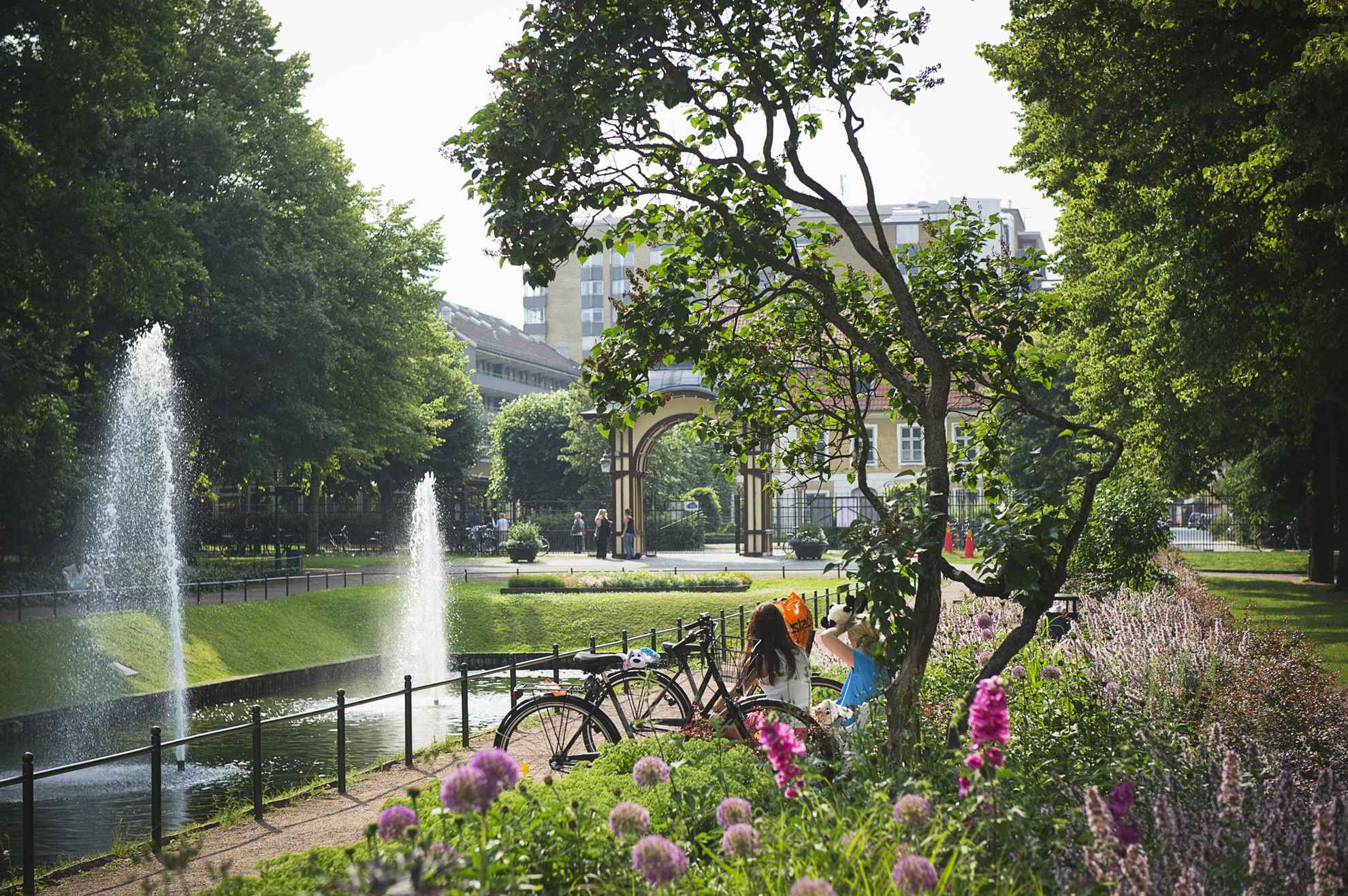 Bloemen en groen in een stadspark. Je ziet de rug van twee vrouwen zitten naast een zwembad met twee fonteinen. Op de achtergrond zie je de ingang en enkele gebouwen buiten het park.