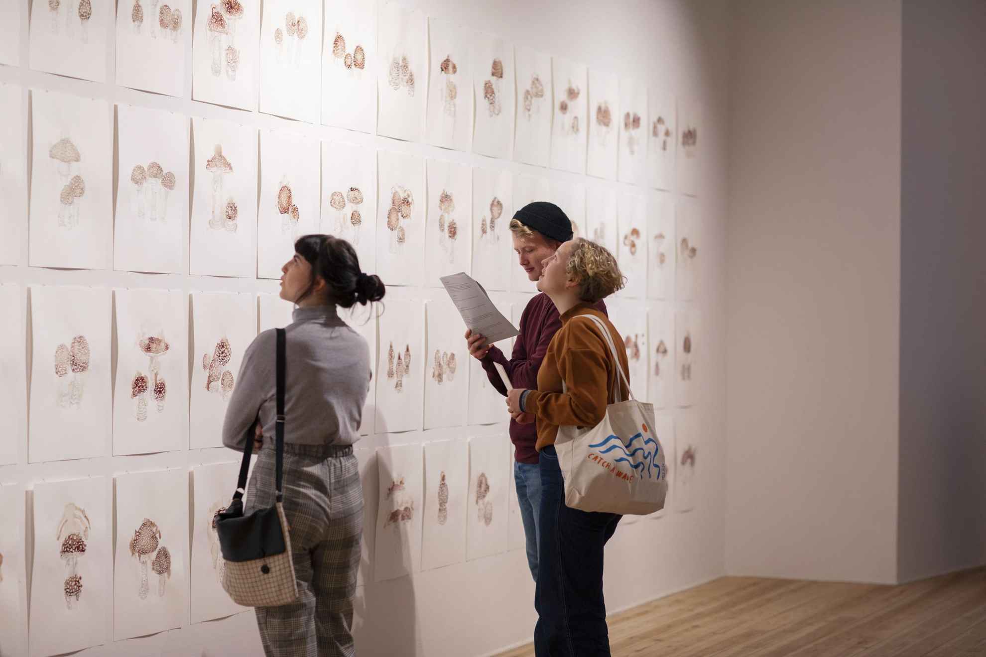 Een man en twee vrouwen bekijken verschillende schilderijen van paddenstoelen op een muur in een museum.