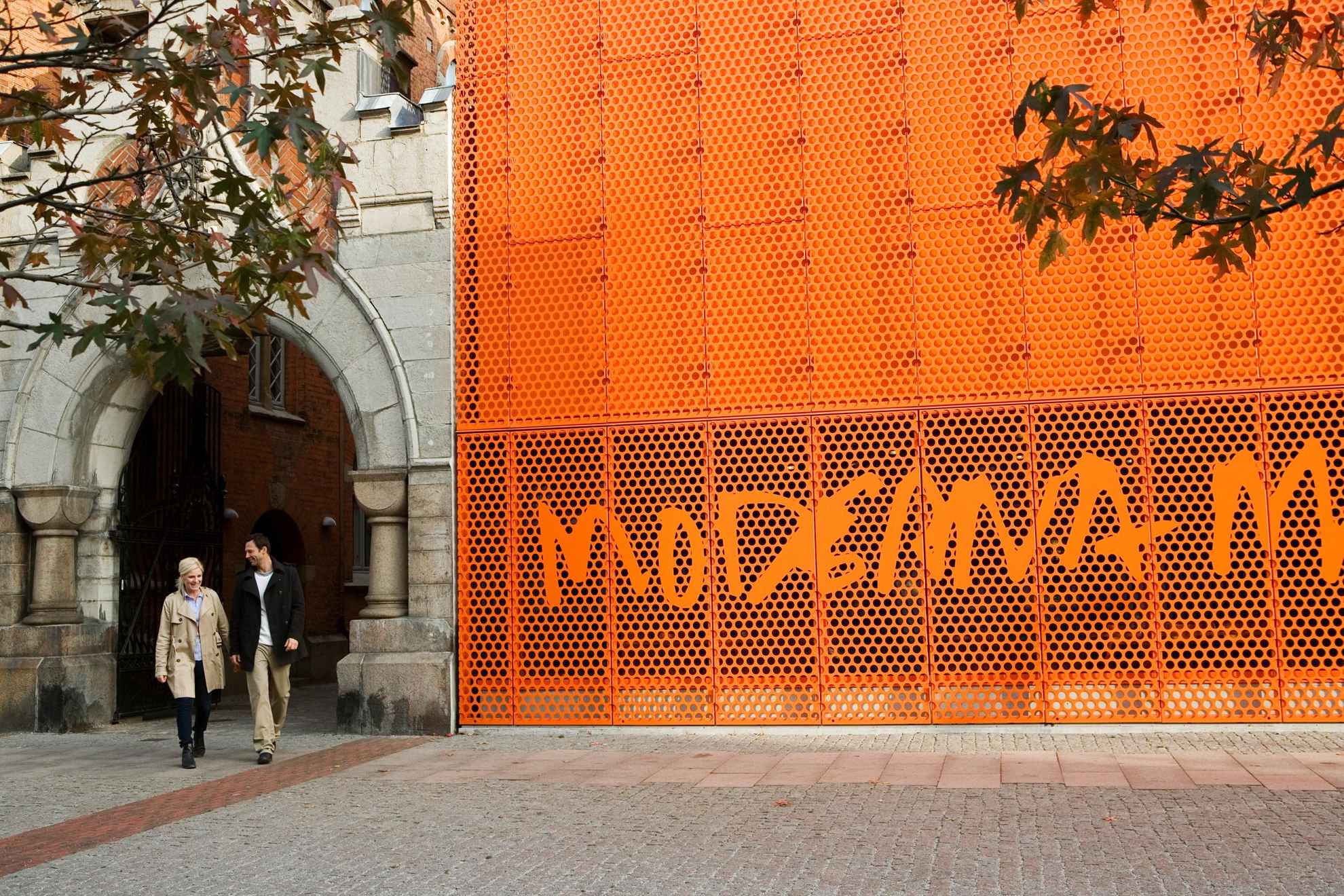 Een man en een vrouw lopen buiten en langs het oranje bord van het Moderna Museet in Malmö.