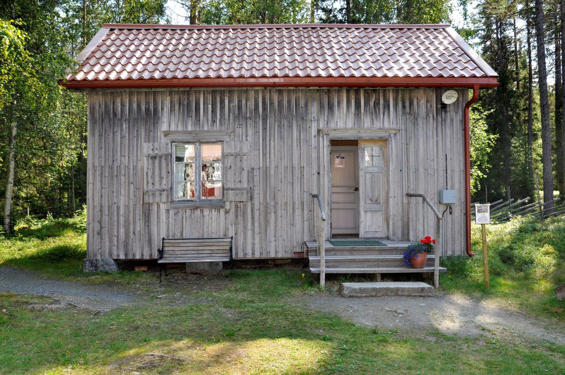 Een eenvoudig, grijs houten huisje met een bloempot bij de ingang, gelegen tegenover een bos.