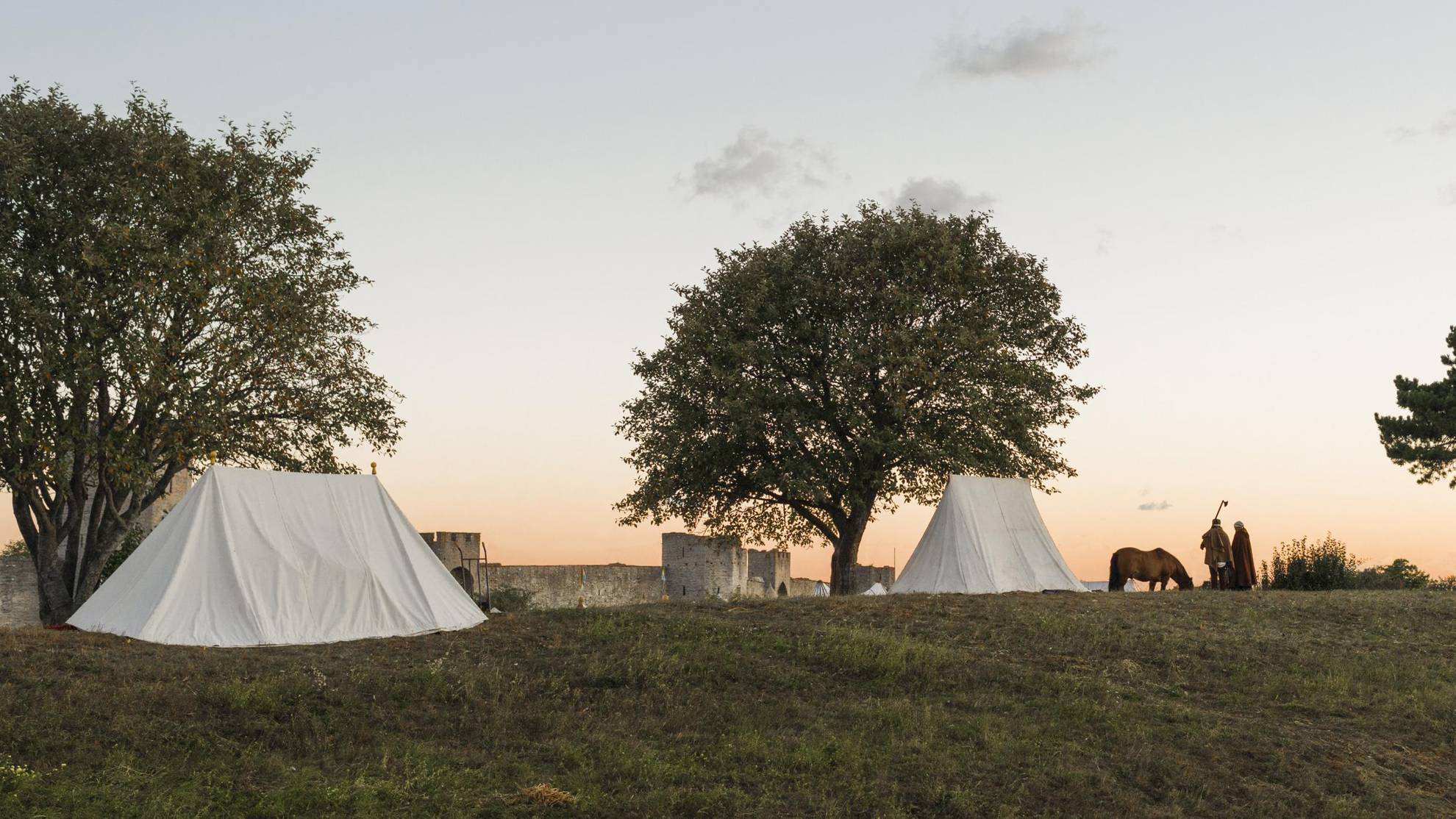 Twee witte tenten naast twee bomen net buiten de stadsmuur van Visby. Twee mensen in middeleeuwse kleding en een paard staan naast een van de tenten.
