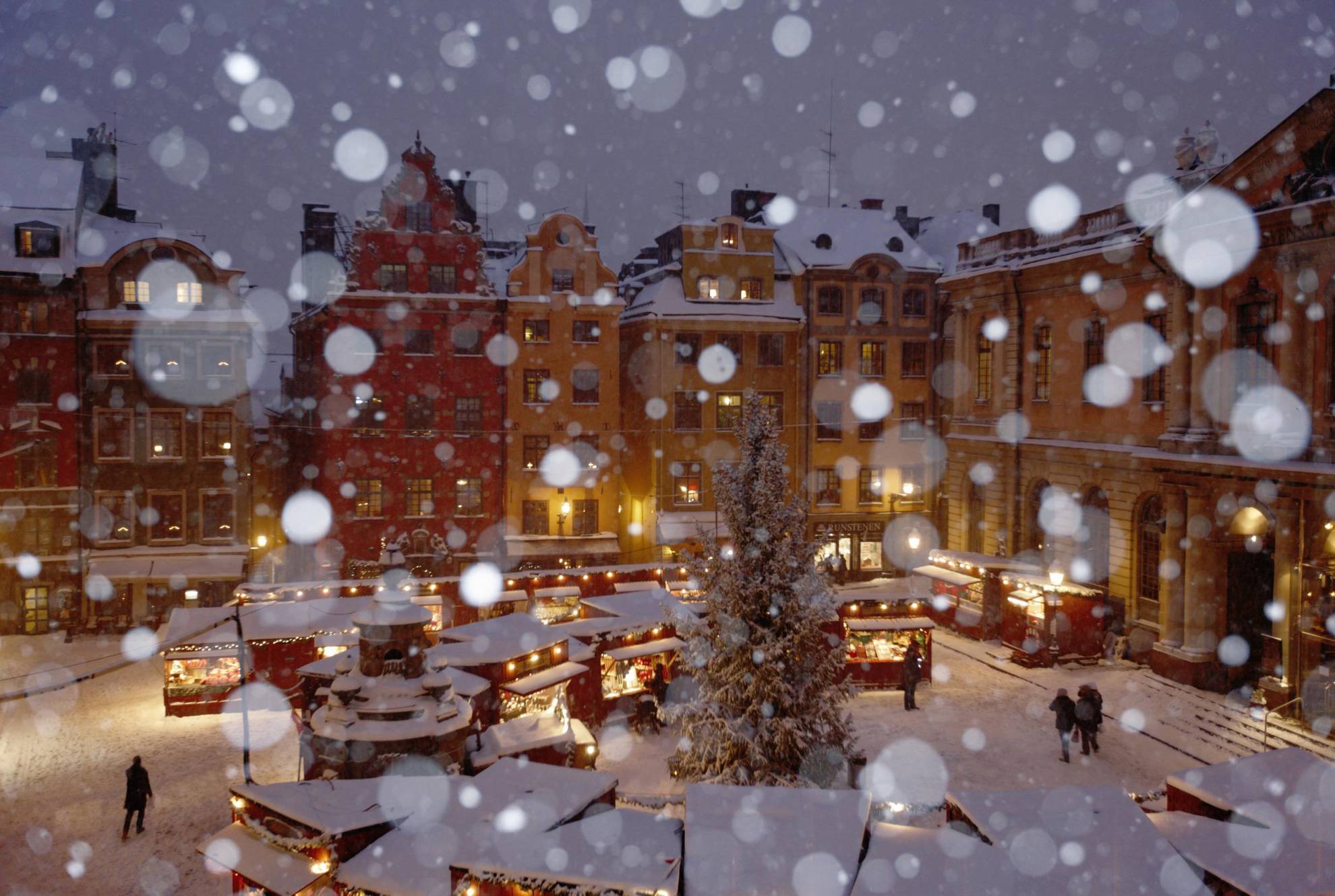 Veel sneeuw met kraampjes op een kerstmarkt voor een rood en een geel huis op een plein.